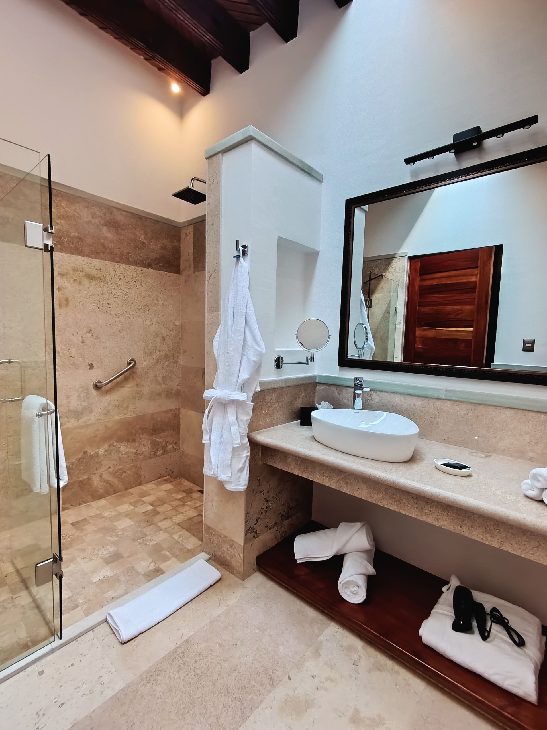 Baño con ducha, tocador y espejo; paredes y suelo de color beige; bata blanca colgada.