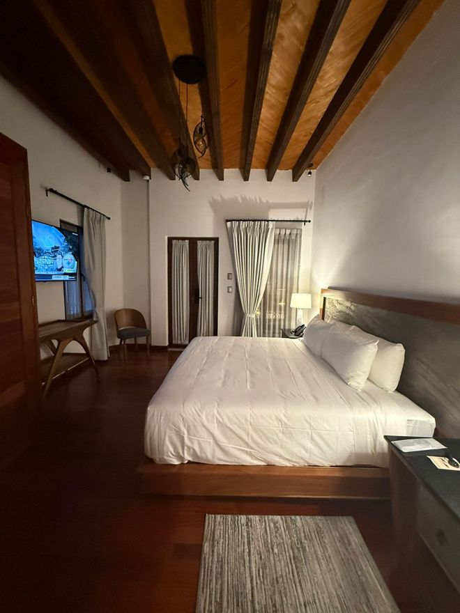 Dormitorio con vigas de madera en el techo, pisos de madera, cama grande, TV y mesa auxiliar.