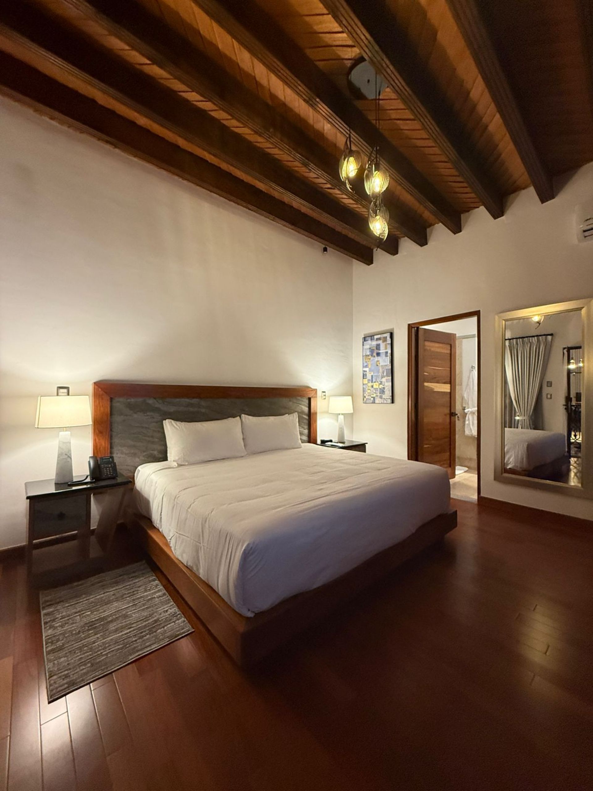Dormitorio con vigas de madera, pisos de madera, cama grande y una puerta que conduce a un baño.