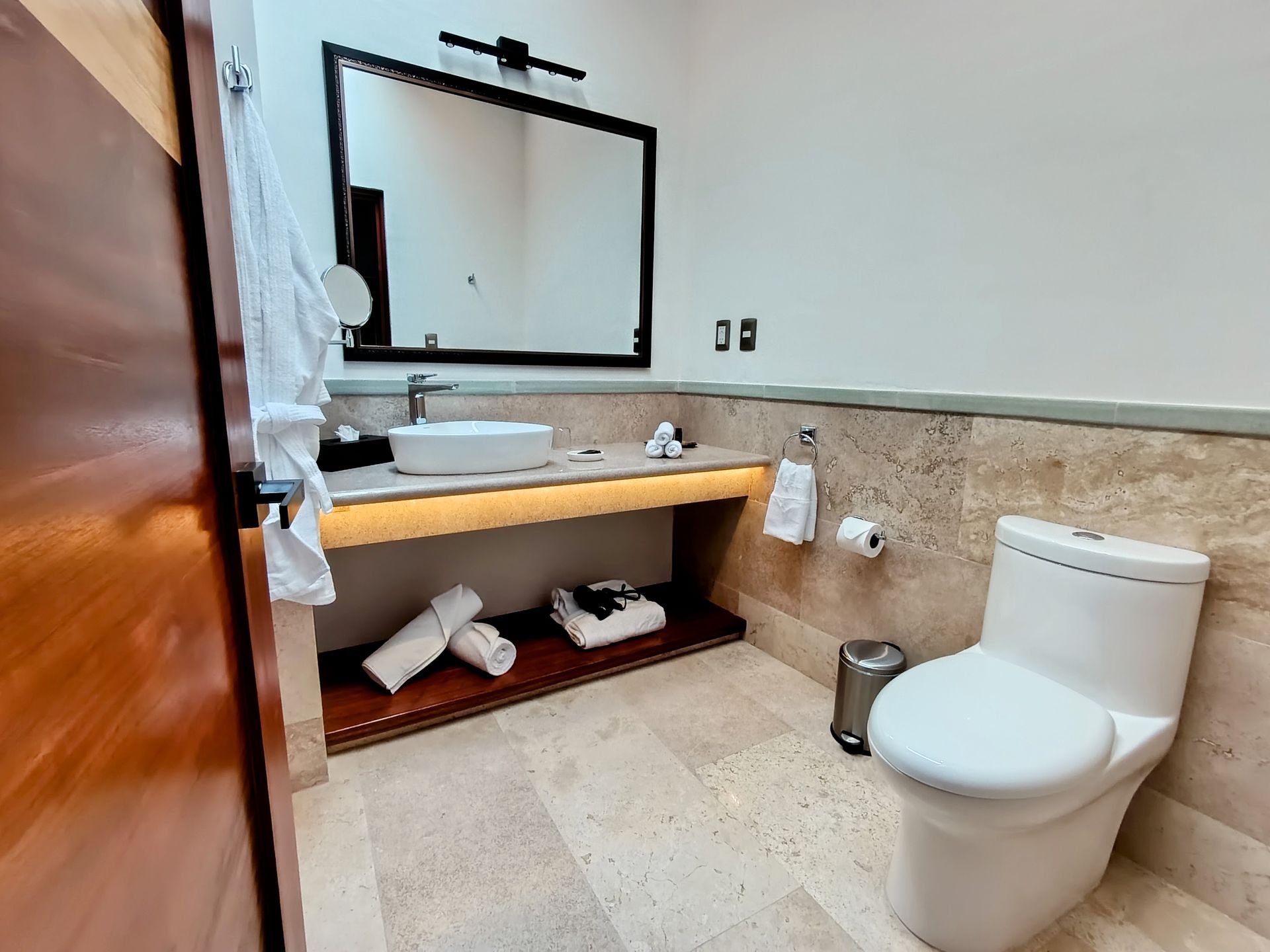Baño con lavabo, espejo, inodoro y toallas enrolladas en un estante. Paredes y suelo de piedra clara.