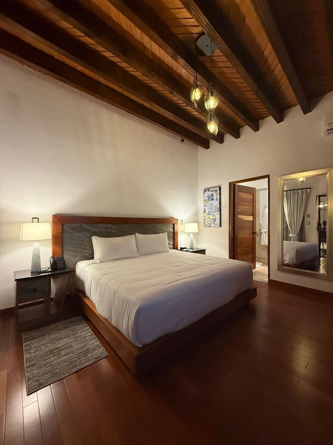 Dormitorio con vigas de madera, pisos de madera, cama grande y una puerta que conduce a un baño.