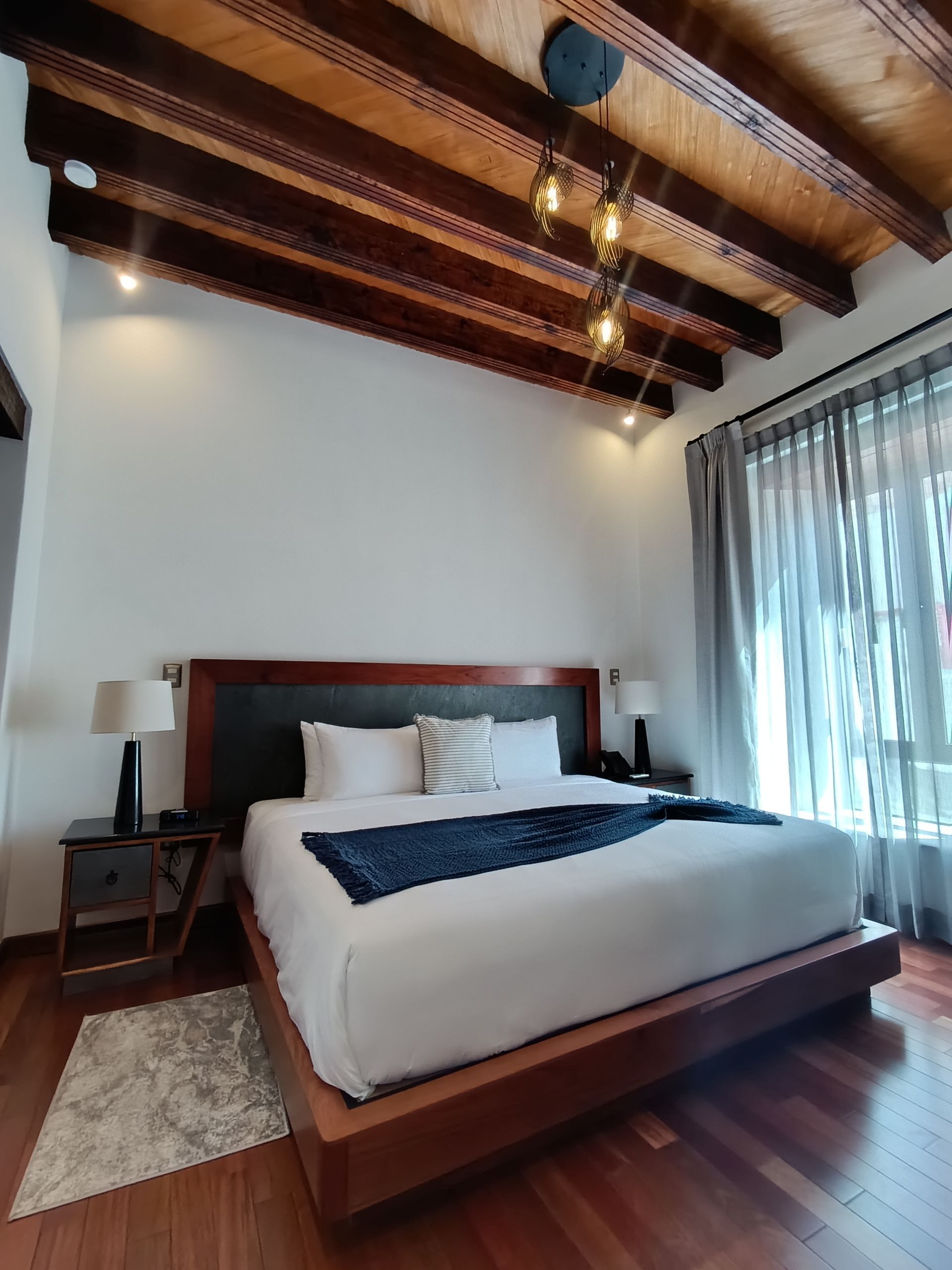 Dormitorio con estructura de cama de madera, vigas, ropa de cama blanca, manta azul oscuro, mesitas de noche y una lámpara moderna.