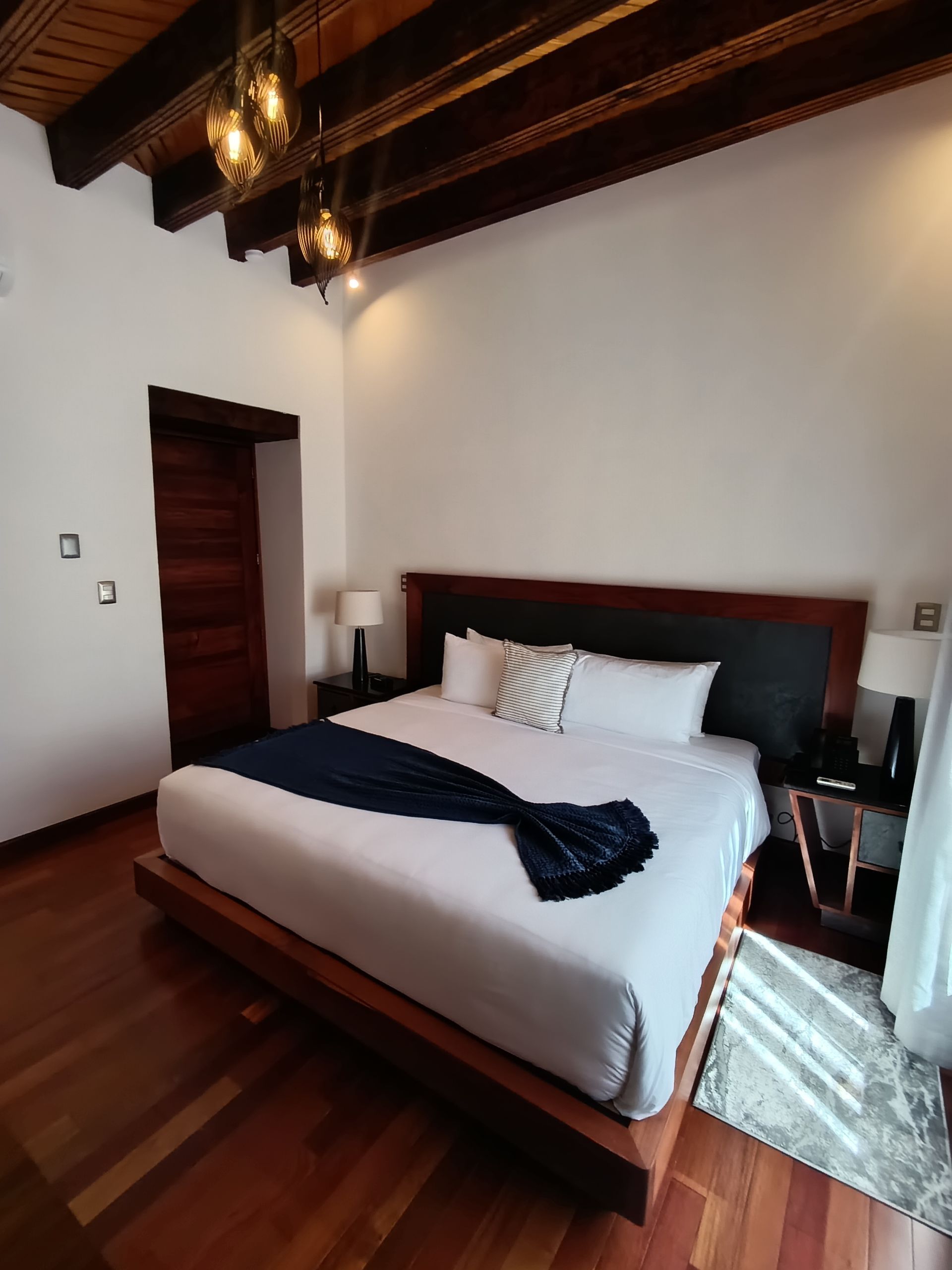 Dormitorio con cama de madera, ropa de cama blanca, manta azul marino, vigas de madera oscura en el techo y piso de madera.