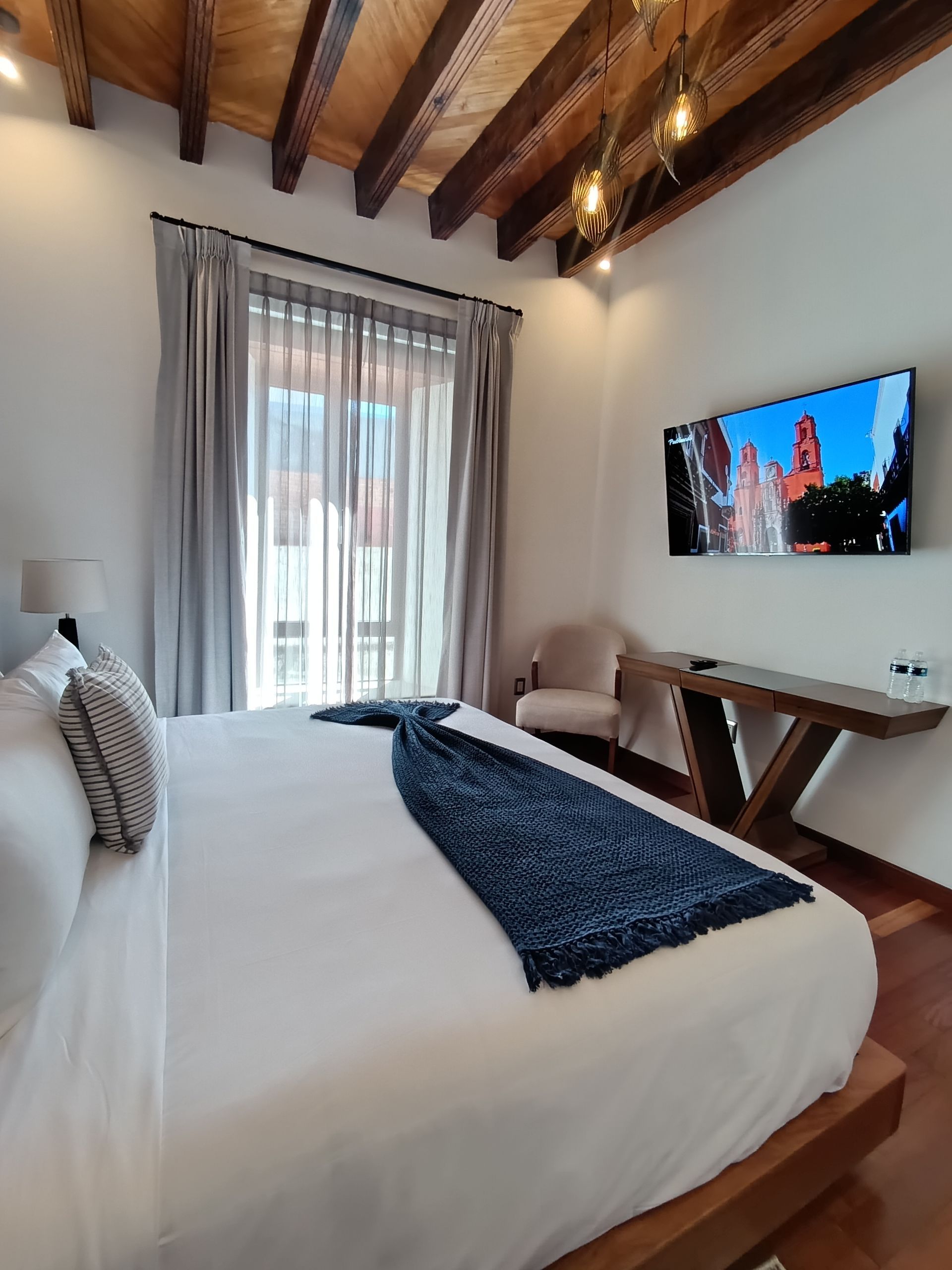 Dormitorio con cama grande, vigas de madera, TV, ventana con cortinas y mesa auxiliar.