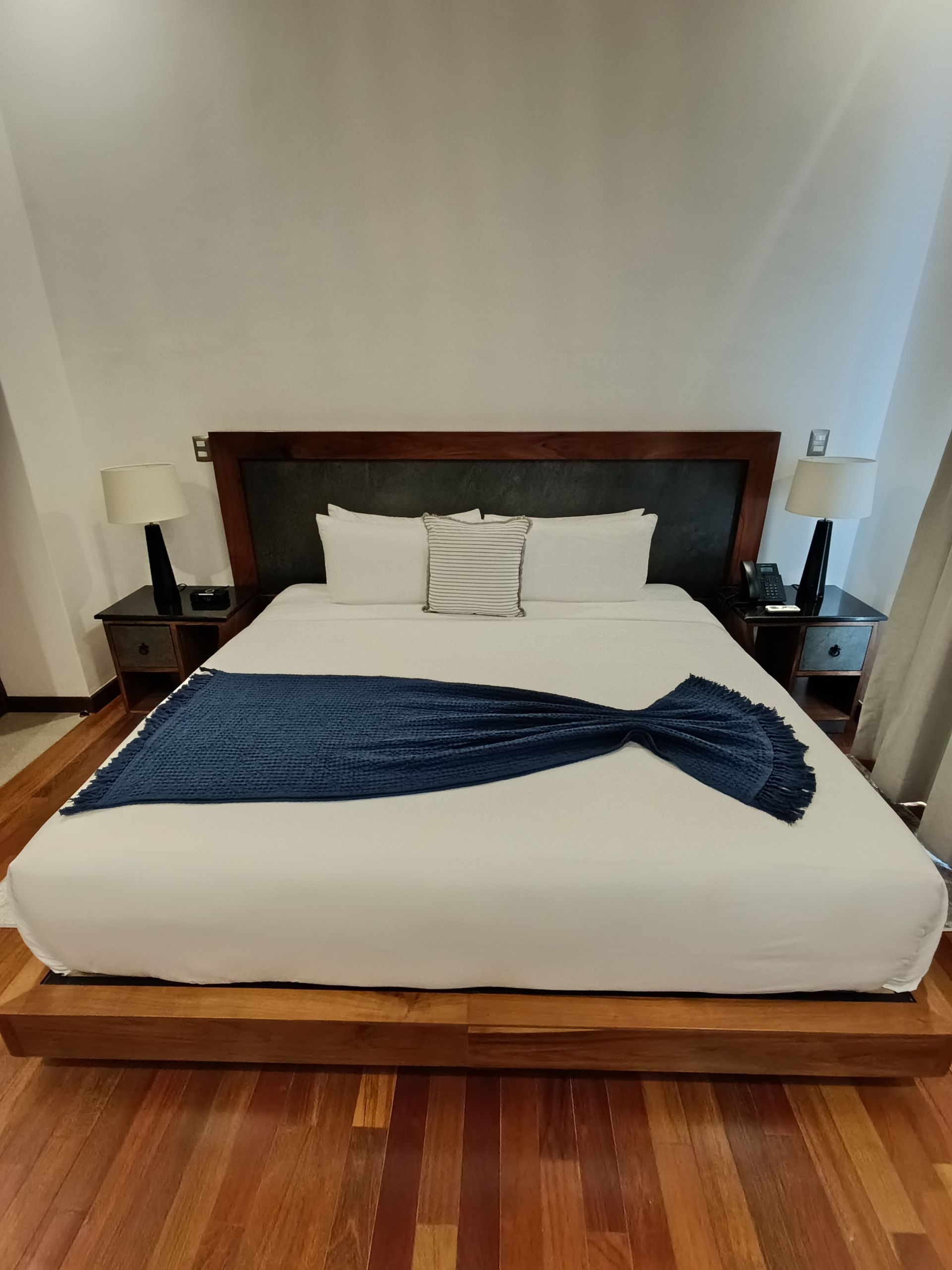 Dormitorio con cama king size. Ropa de cama blanca, manta azul marino, cabecero de madera oscura y mesitas de noche con lámparas. Suelos de madera.