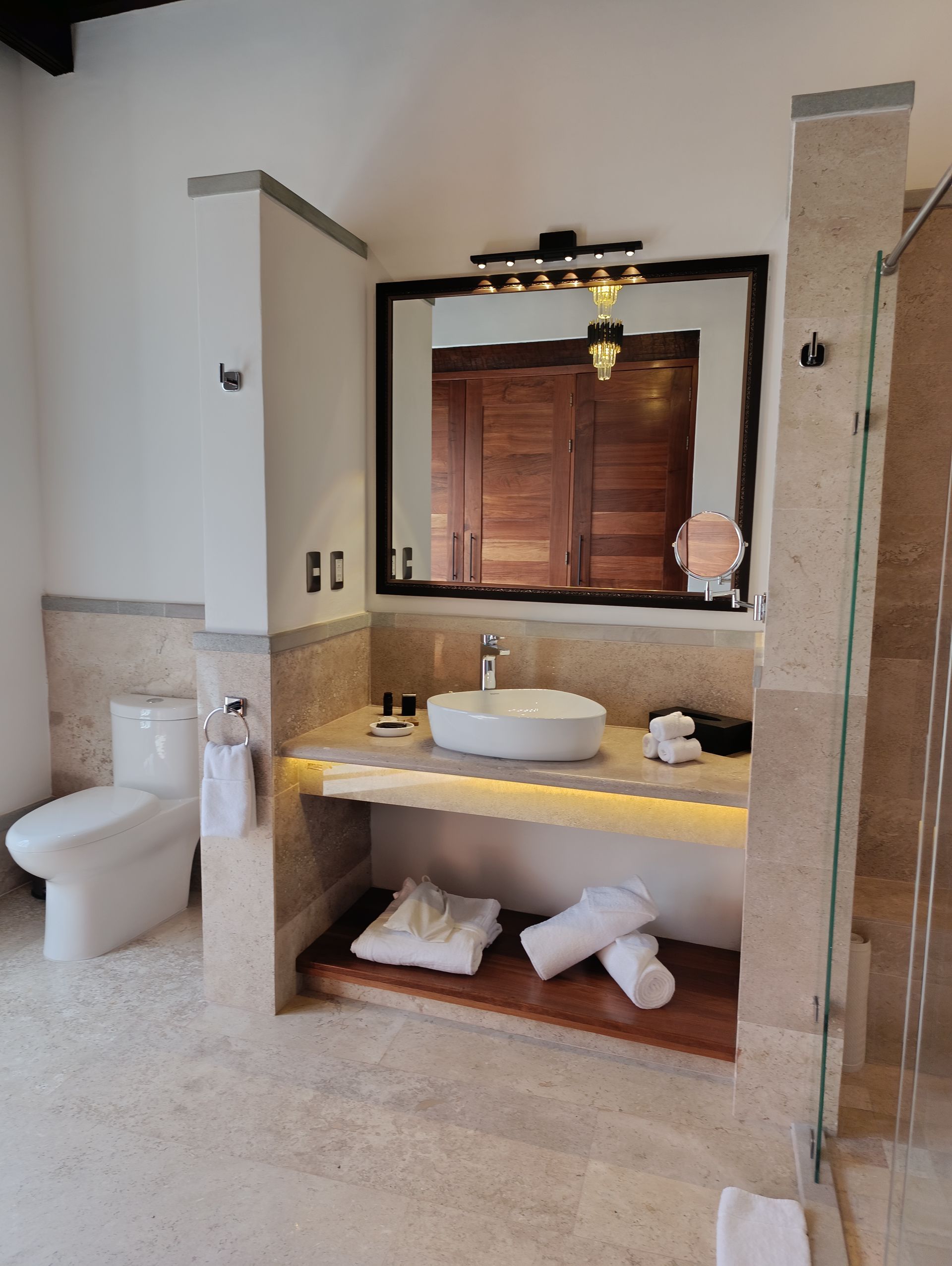 Baño con inodoro blanco, tocador y espejo; toallas en un estante de madera.
