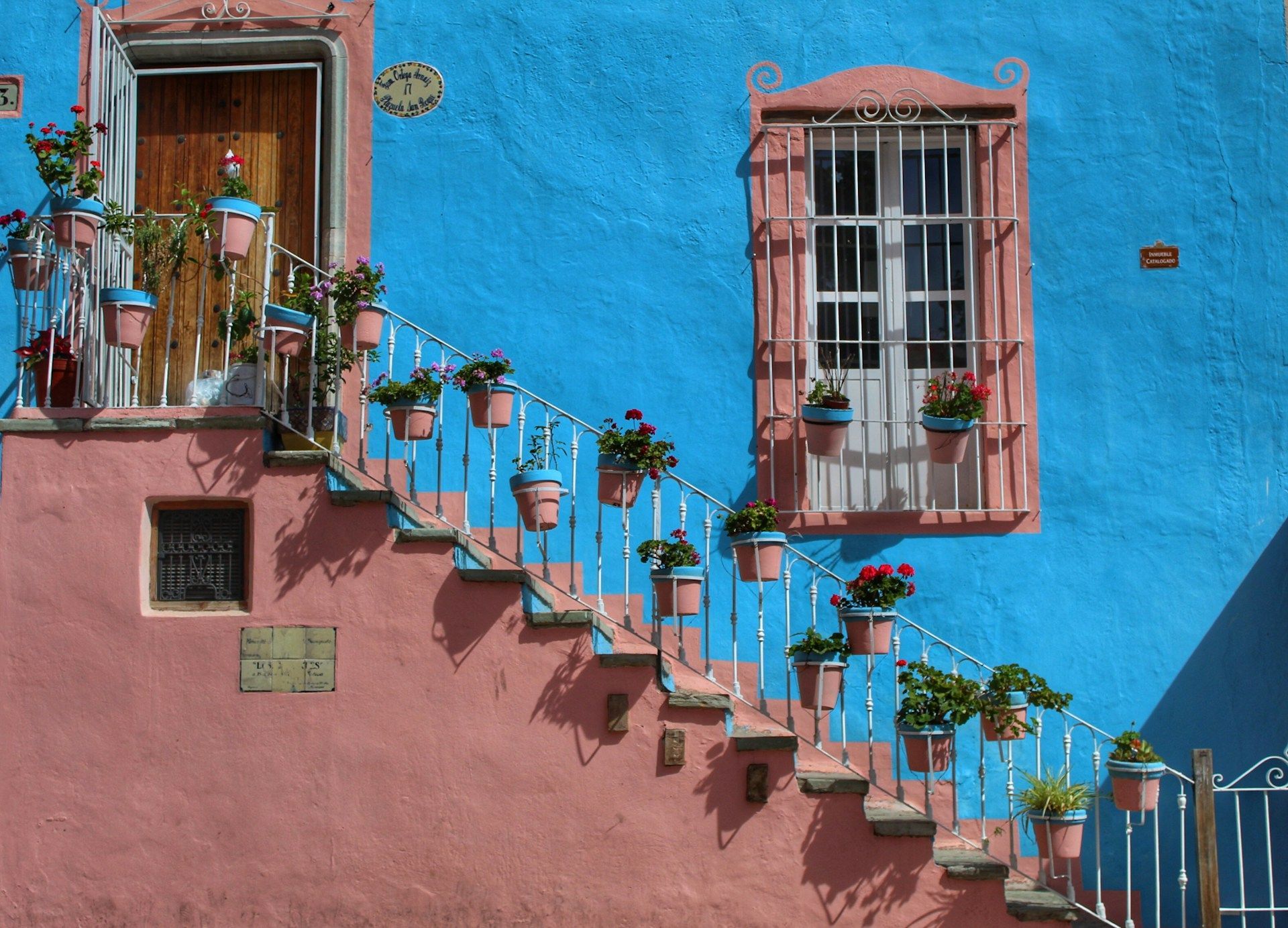 Fachada de edificio colorida con escaleras exteriores adornadas con plantas en macetas; pared azul, borde rosa.