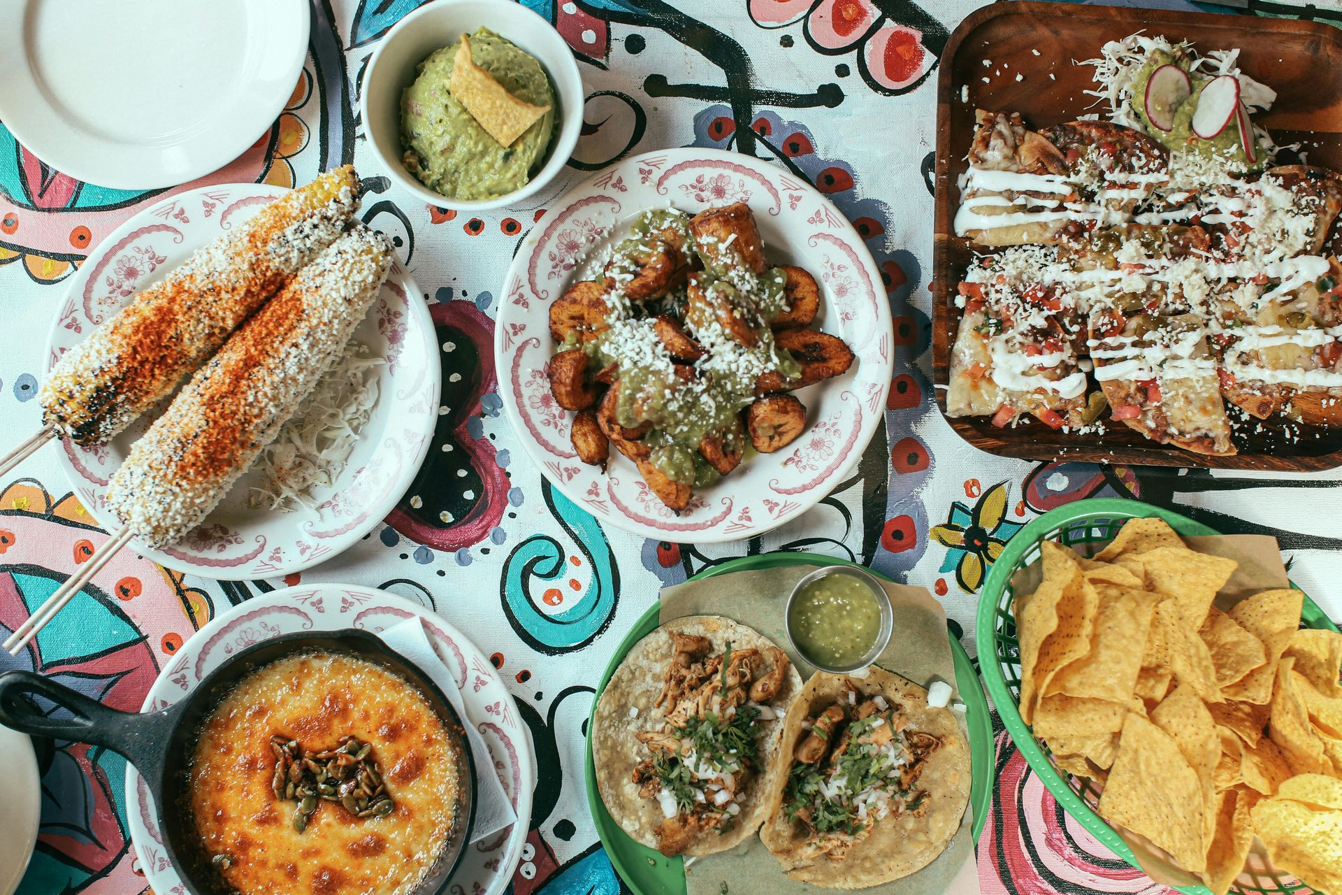 Vista aérea de una mesa colorida con platos mexicanos: elote, tacos, quesadillas, totopos y queso.