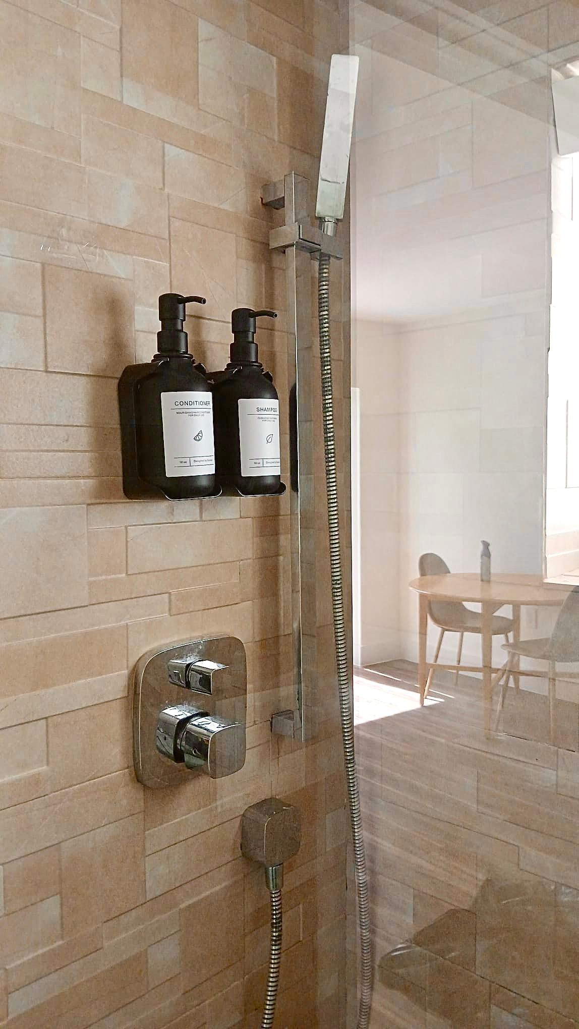 Shower with black soap dispensers on tan tiled wall. Chrome shower head and controls, glass door, view to a dining table.