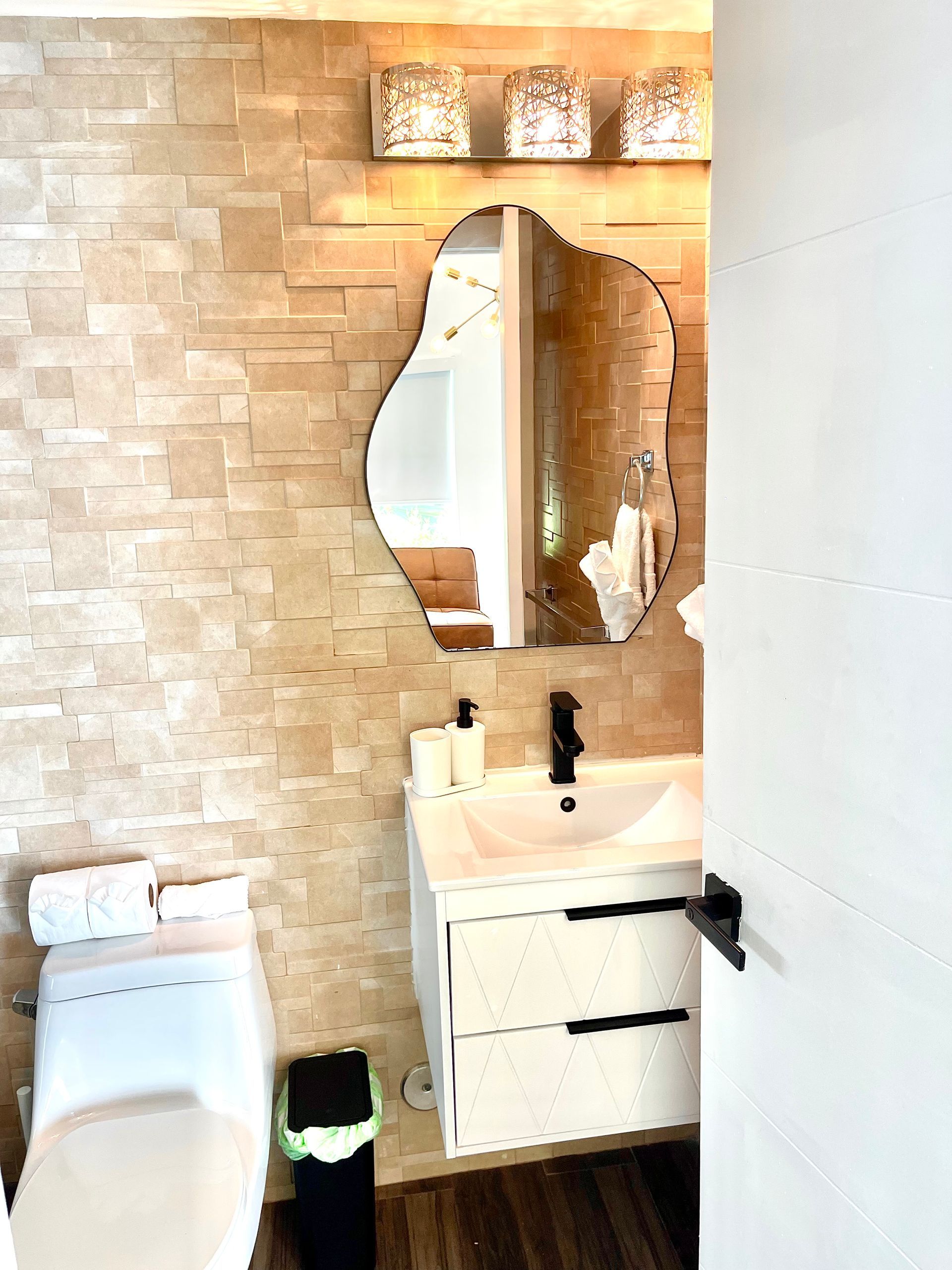 Bathroom with textured beige wall, organic mirror, white vanity, toilet, black accents.