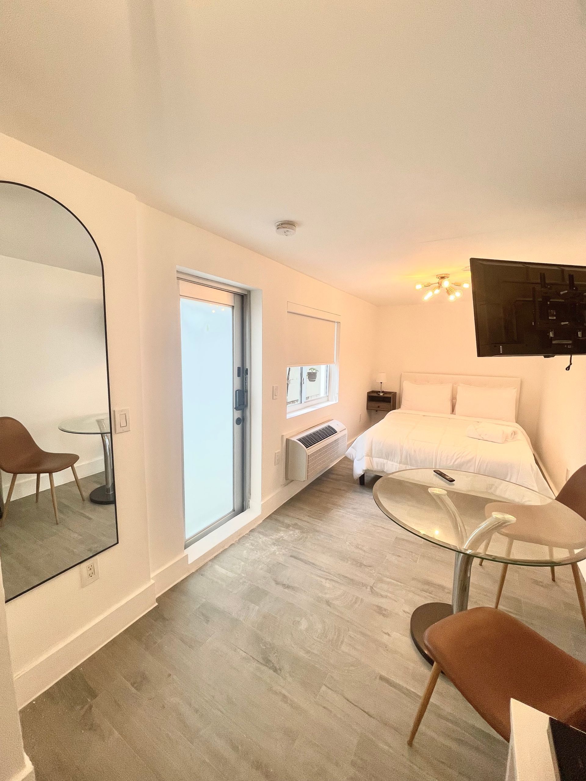 Small, white-walled studio apartment with a bed, TV, small table, mirror, and sliding glass door.