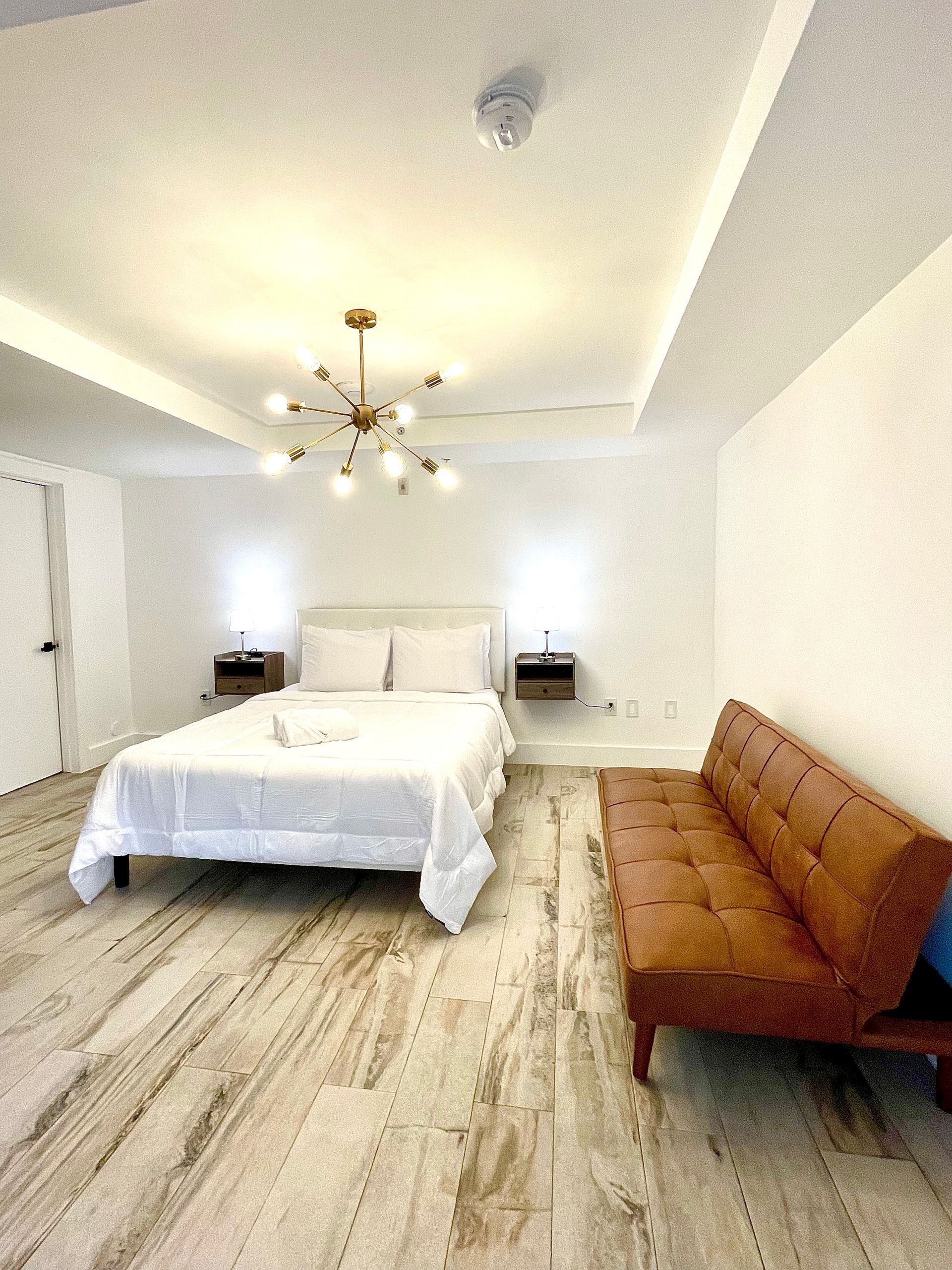 Bedroom with white walls, hardwood floors, bed, brown couch, and gold light fixture.