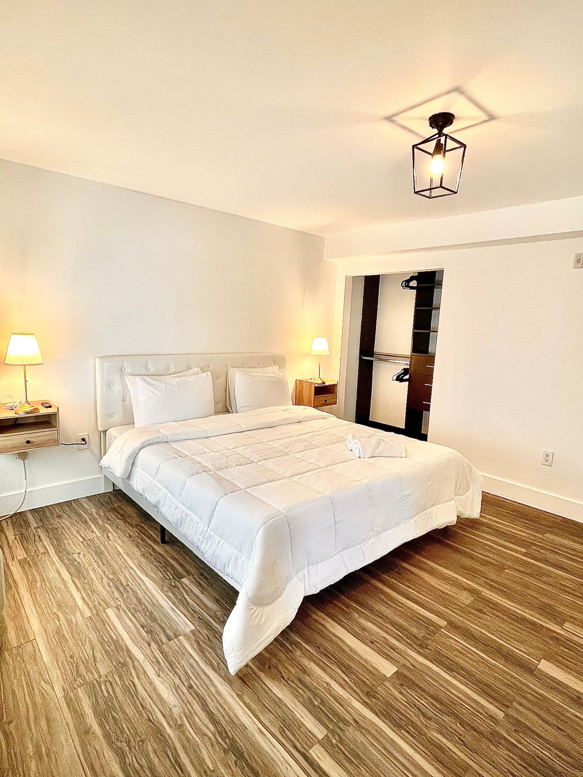 Bedroom with a bed, two nightstands, and open doorway. White bedding, wood floors, and a pendant light.