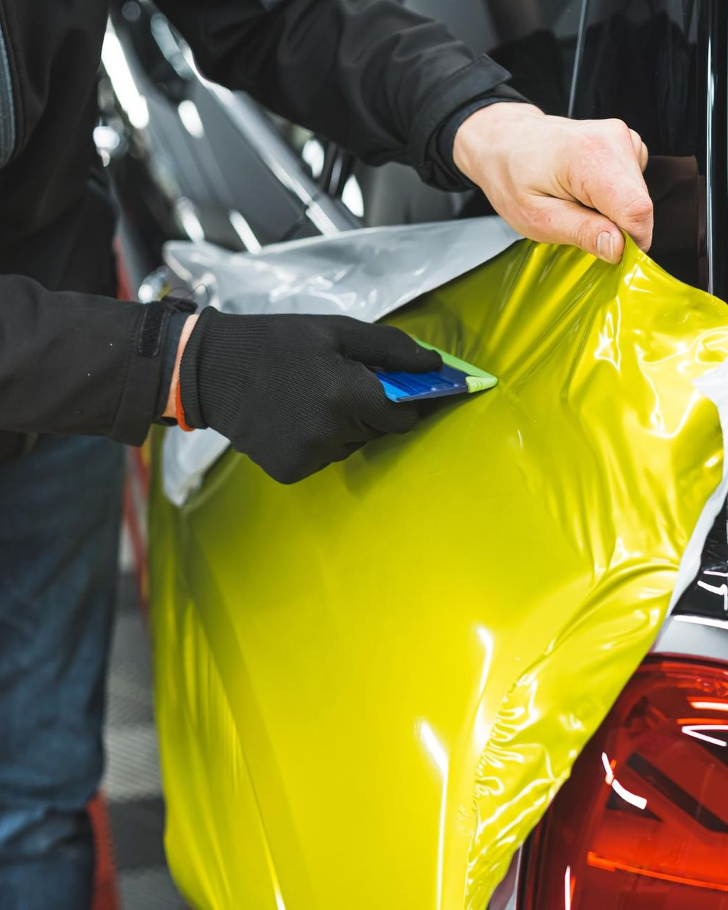 Person in black gloves using a squeegee to apply yellow vinyl wrap to a car's rear panel.