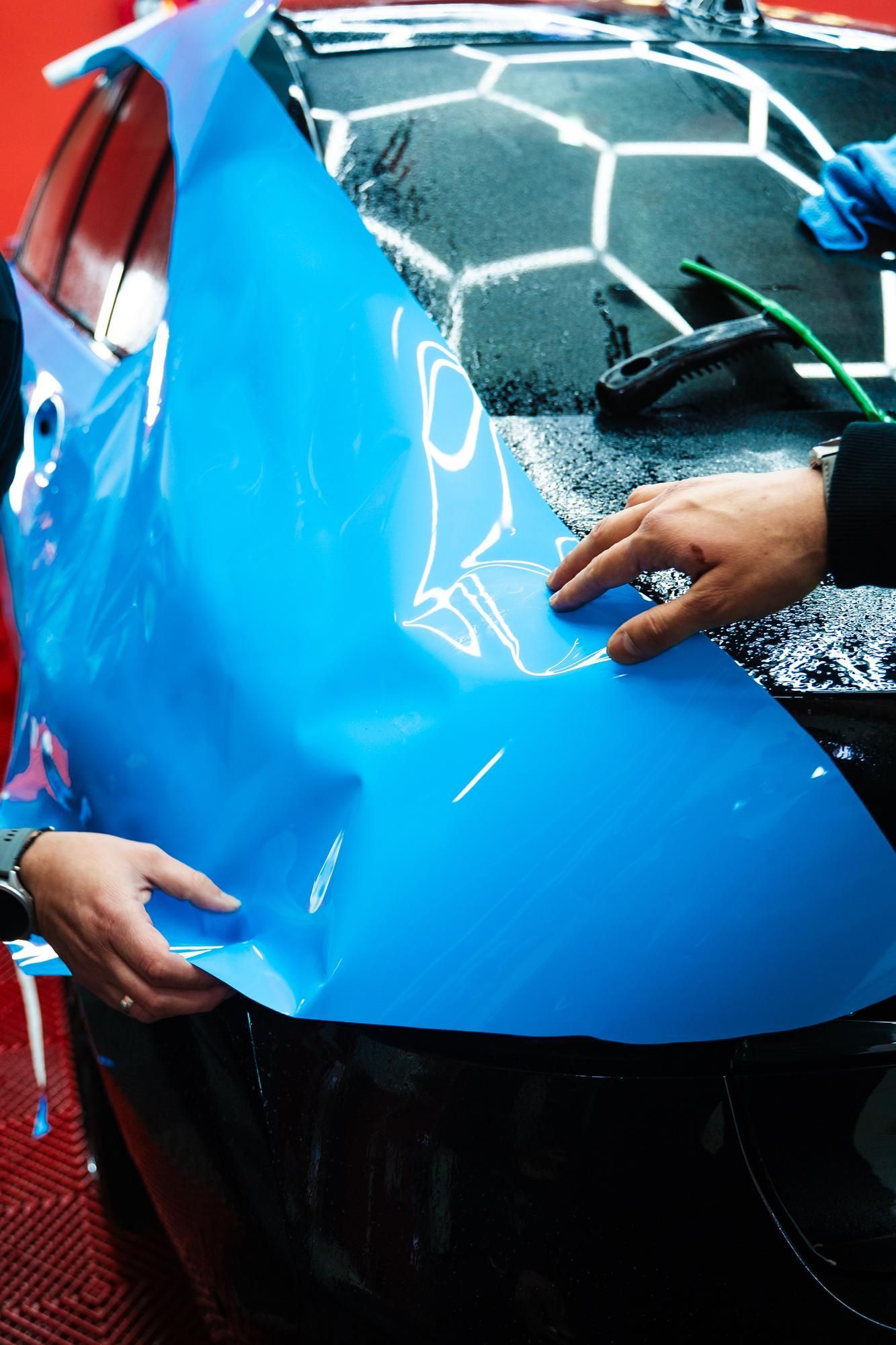 Hands applying blue vinyl wrap to a car's glossy black surface in a well-lit workshop.