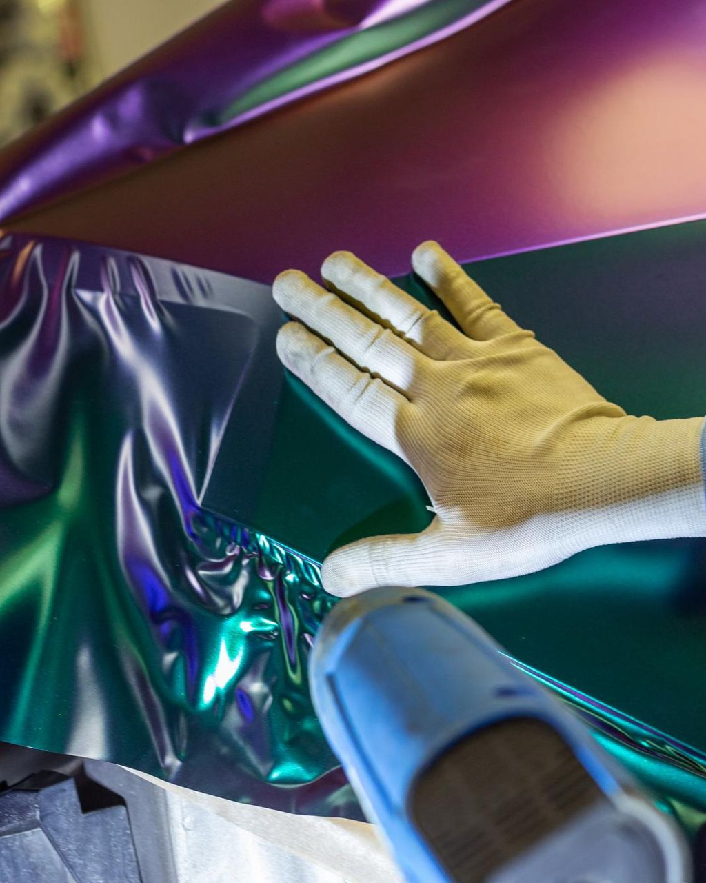 Gloved hand smoothing iridescent vinyl wrap with a heat gun, likely on a vehicle panel, in a workshop setting.
