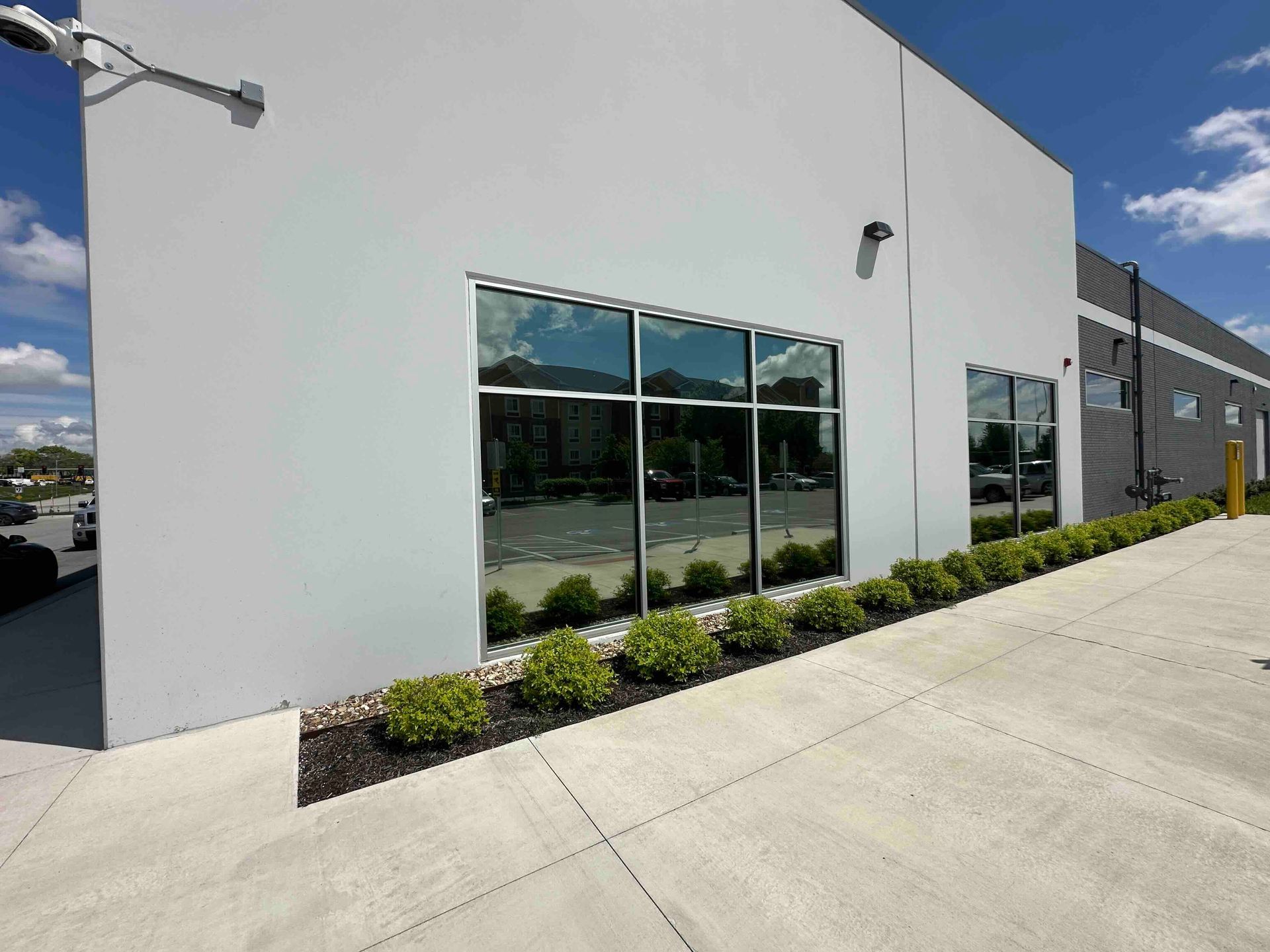 Gray industrial building with reflective windows, lined with shrubs, against a bright blue sky.