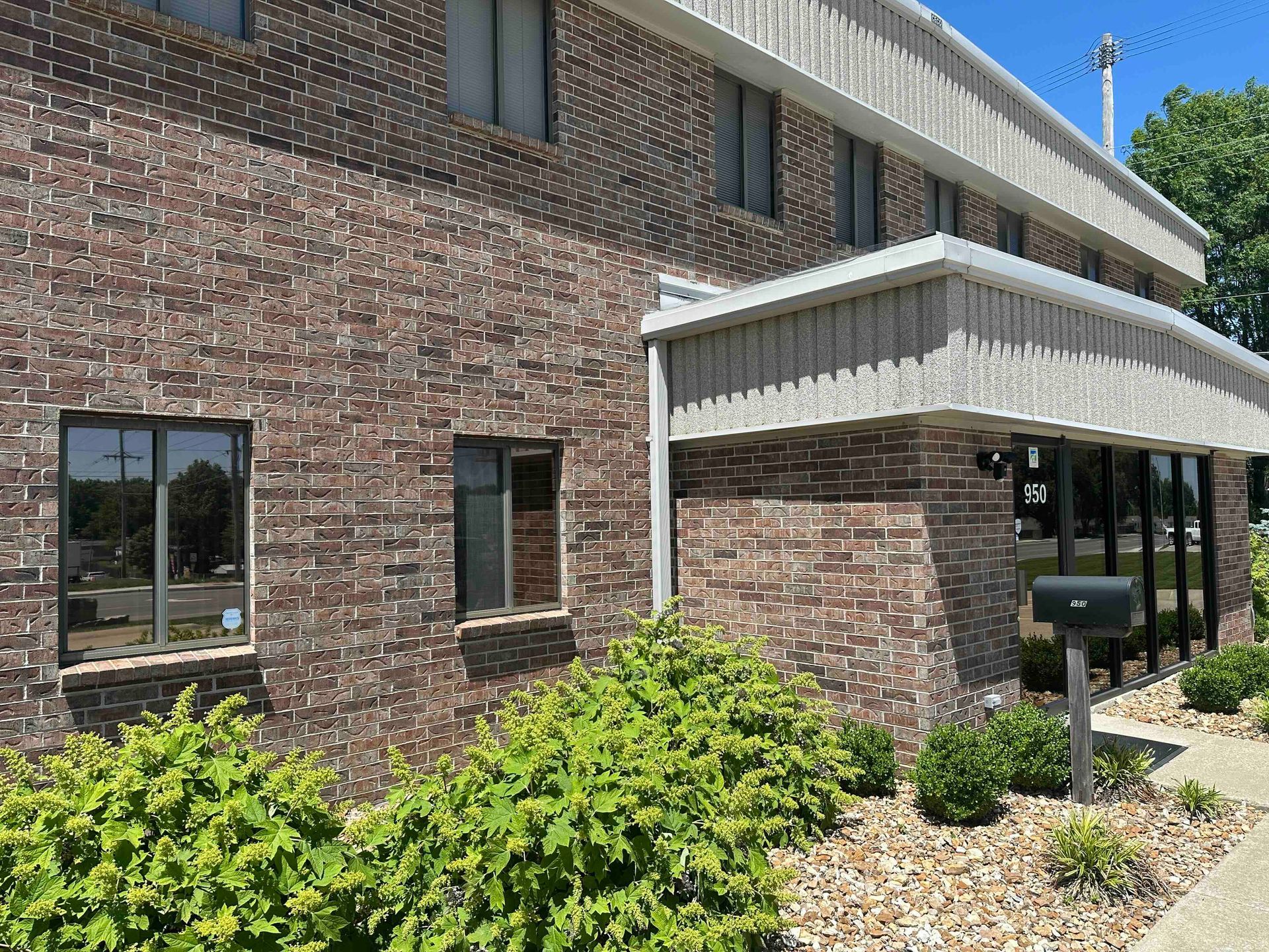 Brick building with multiple windows, small bushes, and a covered entrance.
