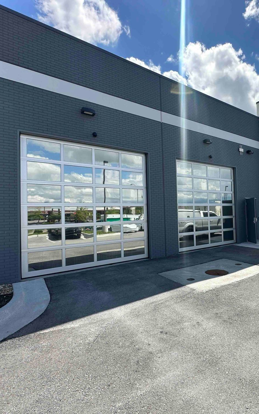 Two gray garage doors with clear, grid-patterned glass, reflecting the sky and vehicles.