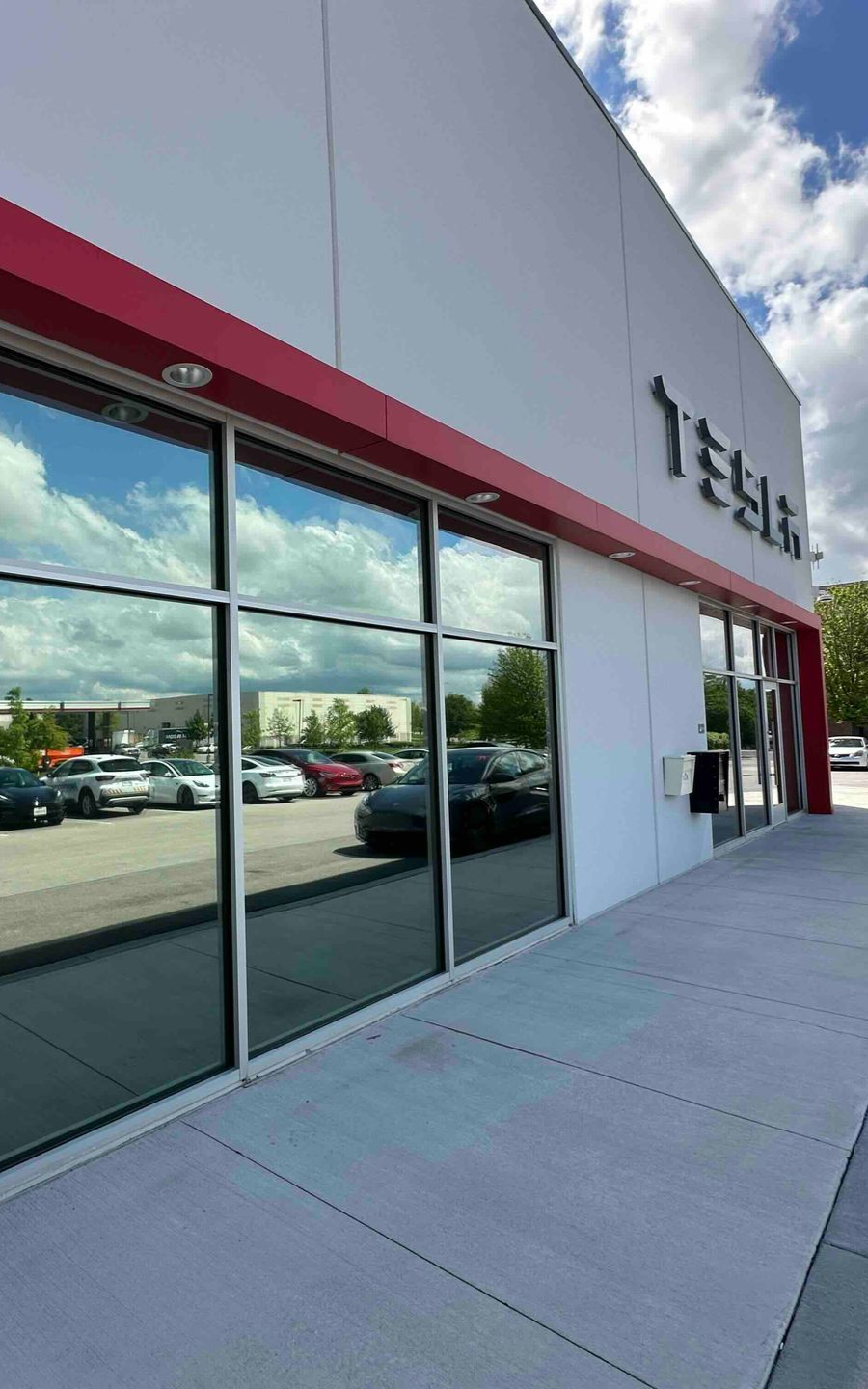 Exterior of Tesla store with reflective windows showing parked cars and sky; red trim and white walls.