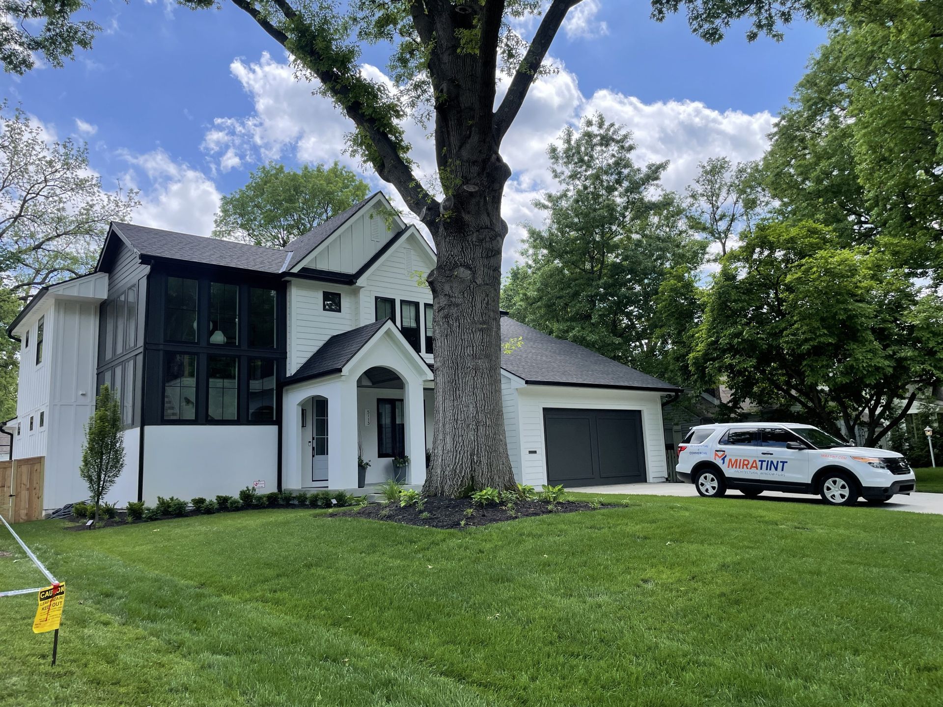 White house with black accents, large tree in front, green lawn, blue sky, and a car in the driveway.