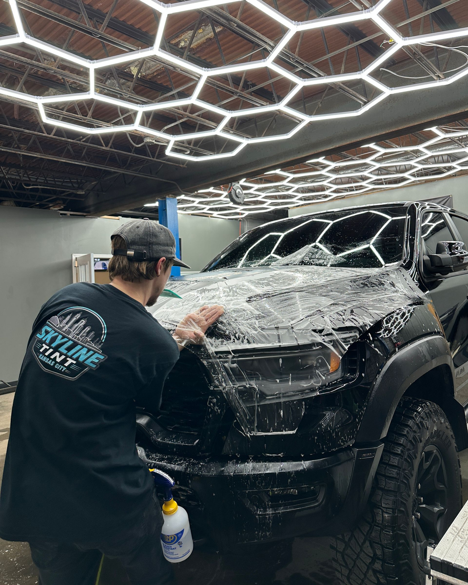 Person applying tint to a car window with a squeegee. The car is blue-gray. Indoor setting.