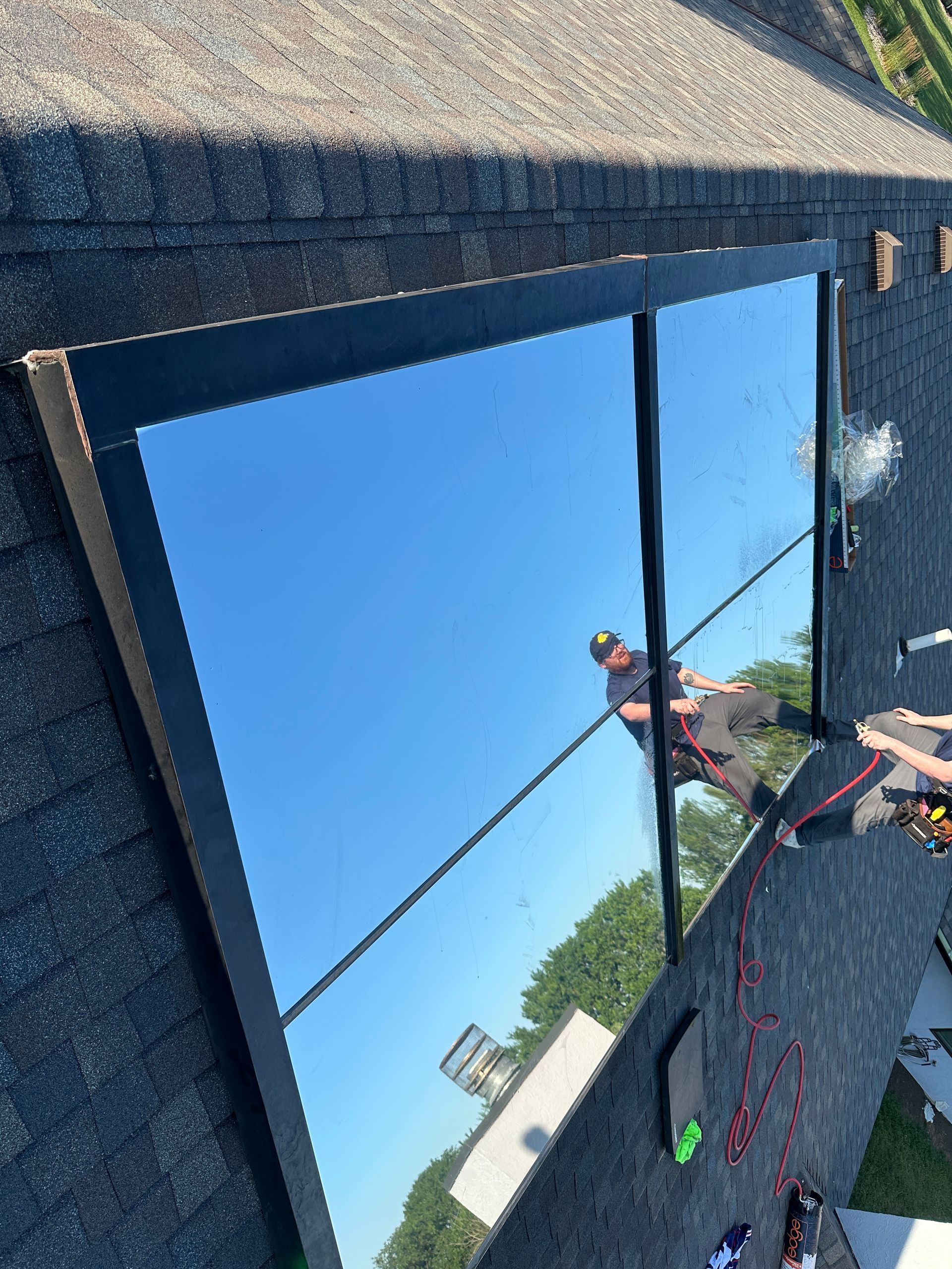 Large mirror on a rooftop reflects a blue sky and person, surrounded by shingles and tools.