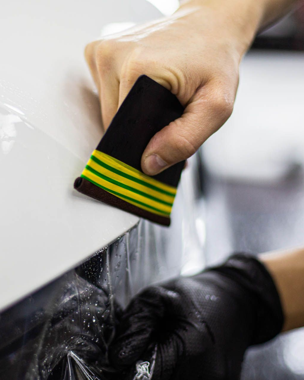 Person using a squeegee to apply clear film to a white car panel, wearing a black glove.