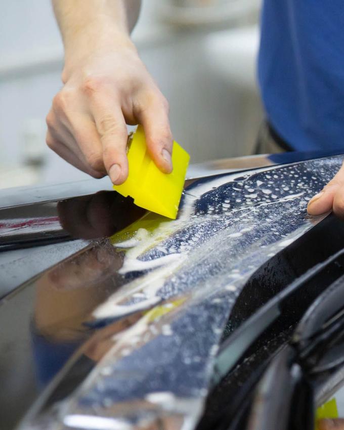 Person applying film to a car surface with a yellow squeegee.