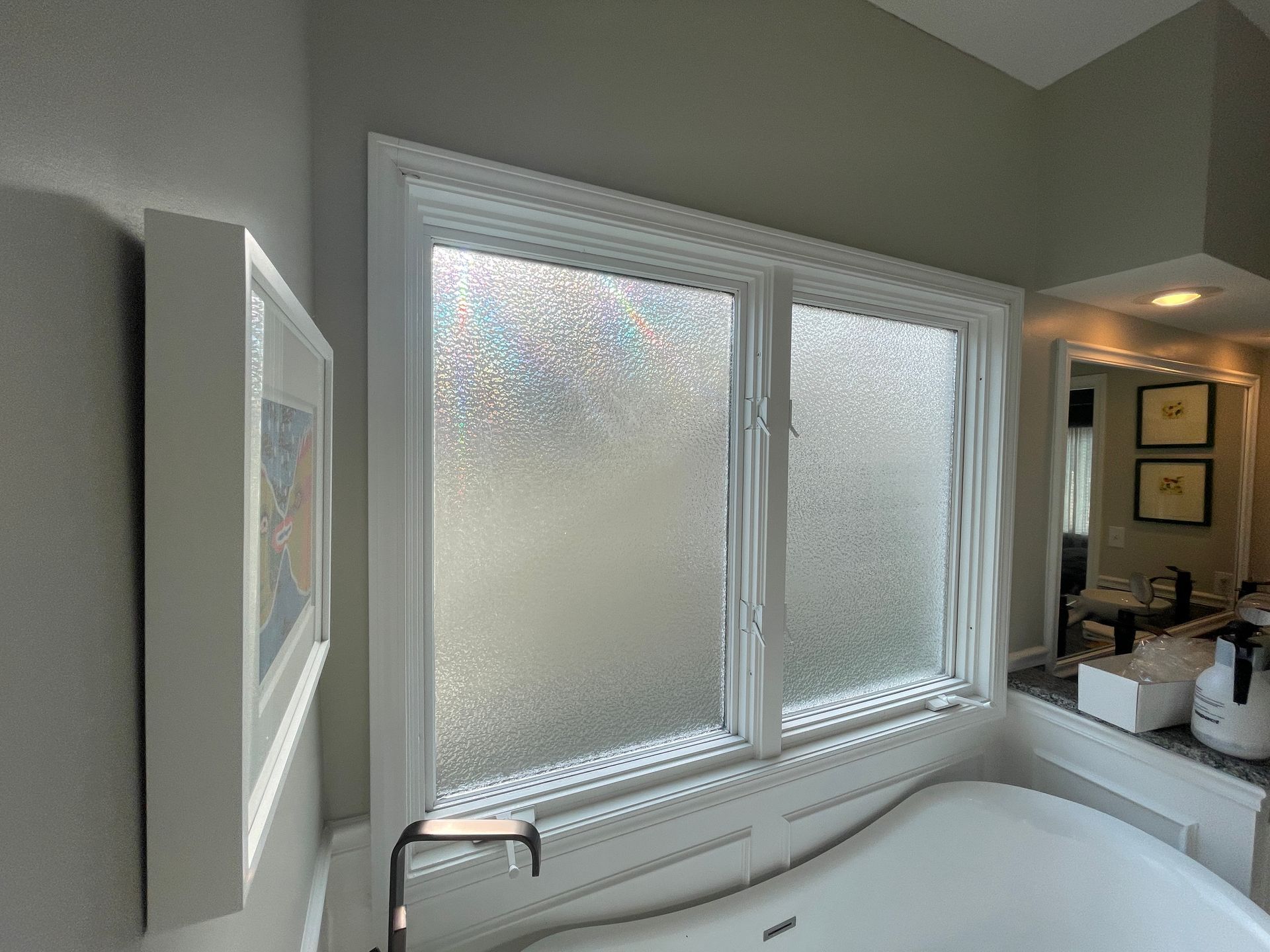 Bathroom window with frosted glass above a bathtub. White frame against gray walls, view of mirror.