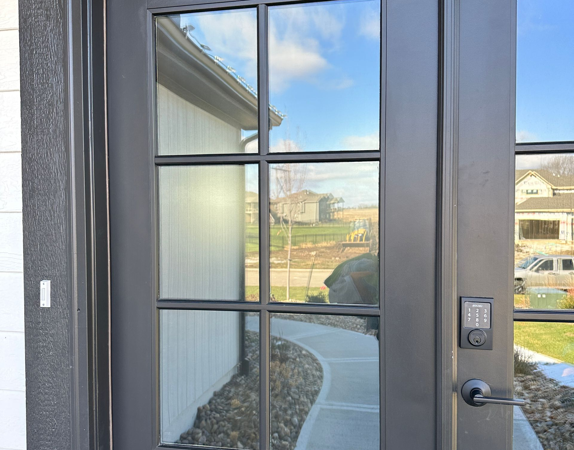 Exterior door with black frame and grid pattern, reflecting a walkway and houses under a blue sky.