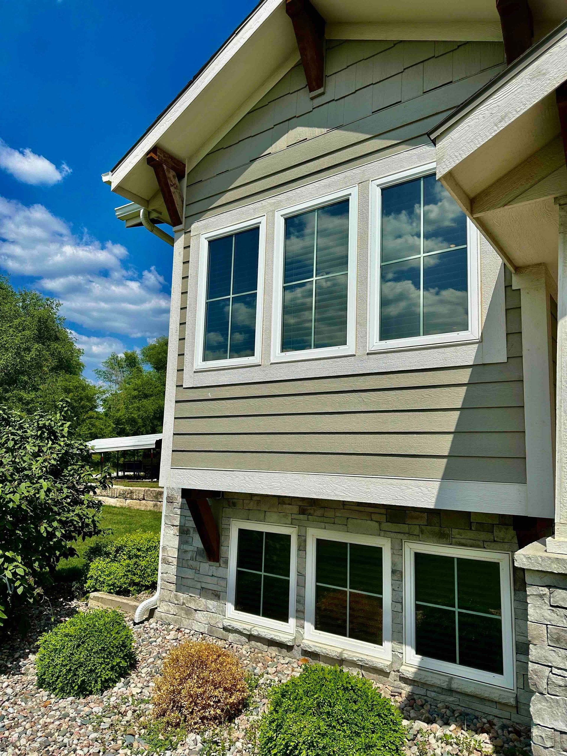Exterior view of a house with gray siding, white trim, and three windows. Blue sky and greenery in the background.