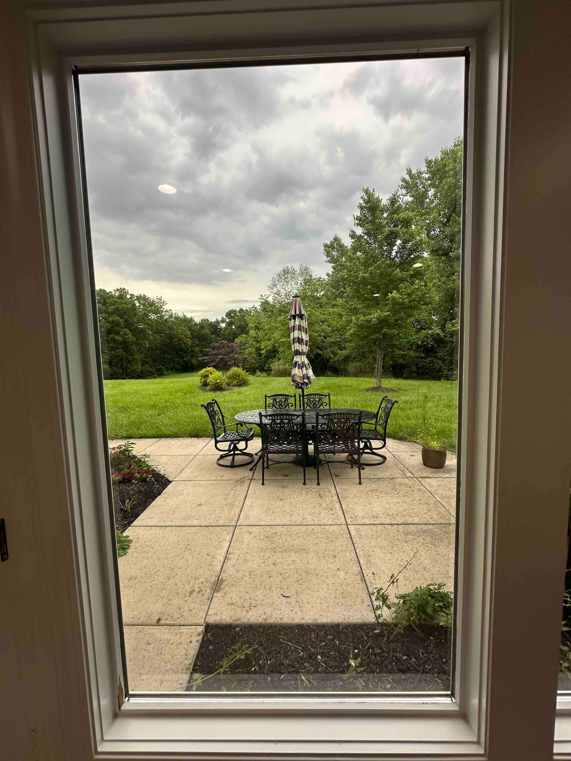 View through a window of a patio with a table, chairs, and umbrella, surrounded by grass and trees under a cloudy sky.