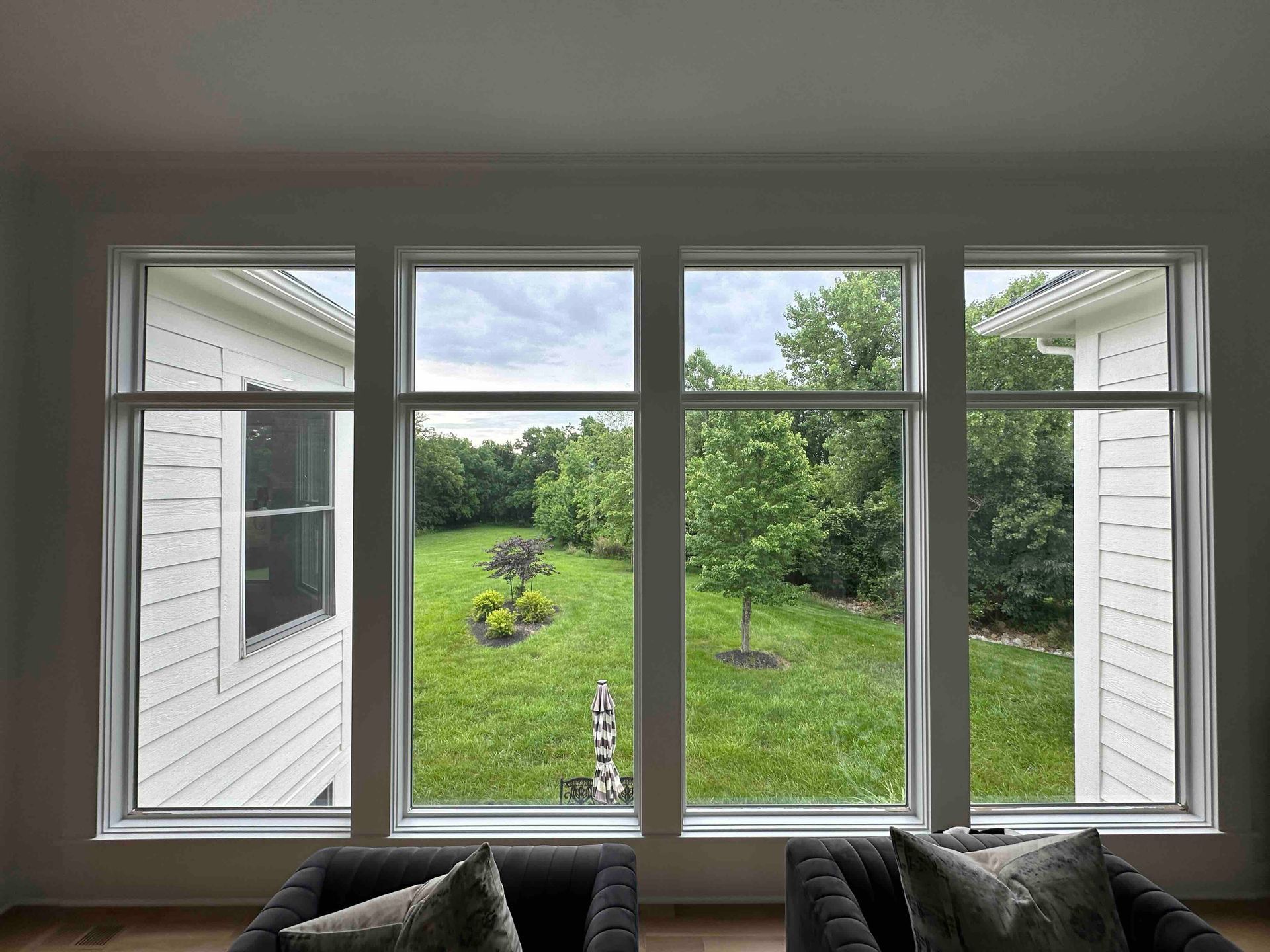 View through a large window to a green backyard with shrubs, trees, and white house siding