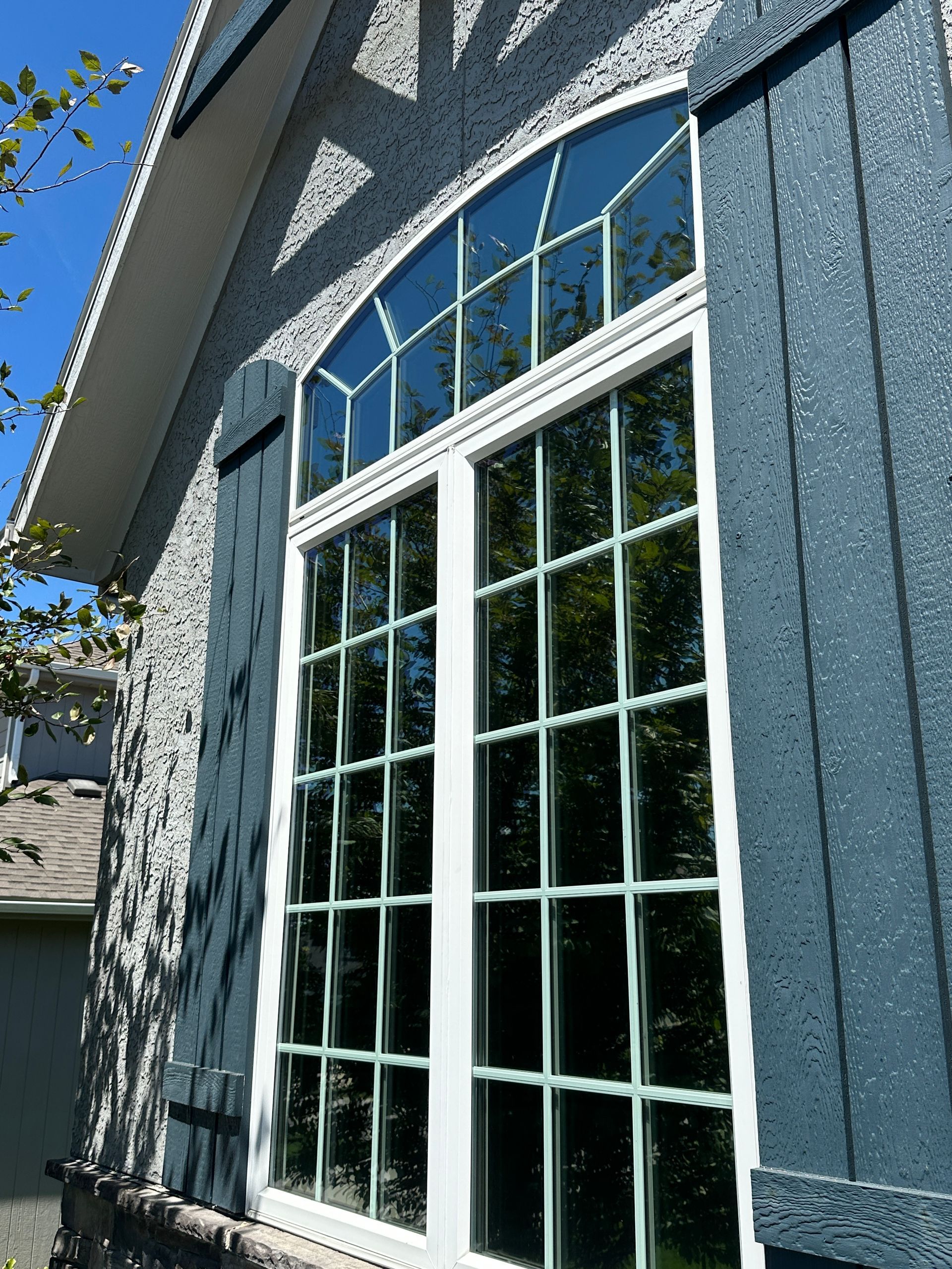 Large, arched window with white trim on a blue stucco house, reflecting trees and sky.