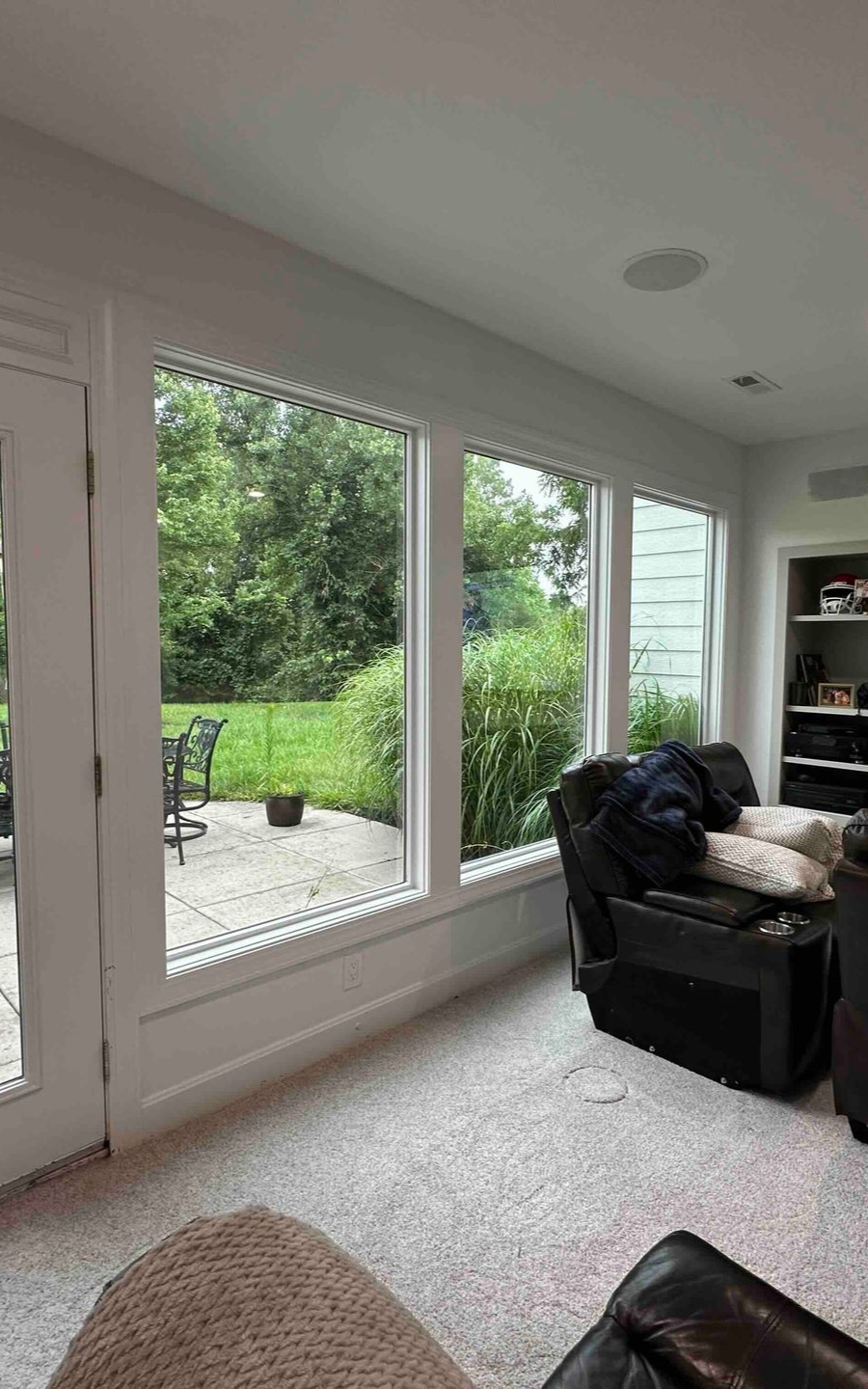 Living room with large windows overlooking a patio and greenery. Black chair and shelves are visible.