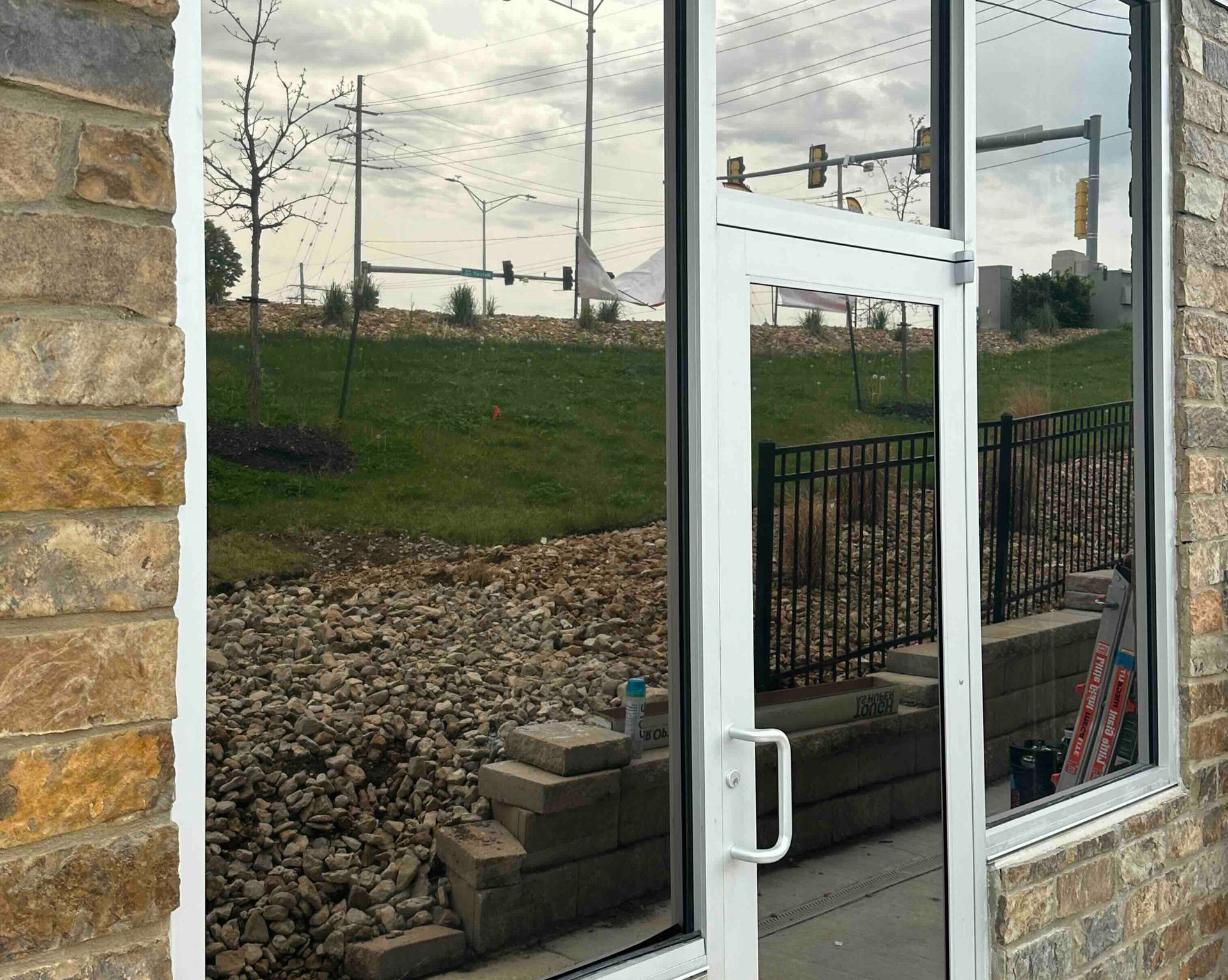 Exterior view: mirrored glass windows and door reflect a landscape with grass, rocks, and a fence. Brick wall on the left.