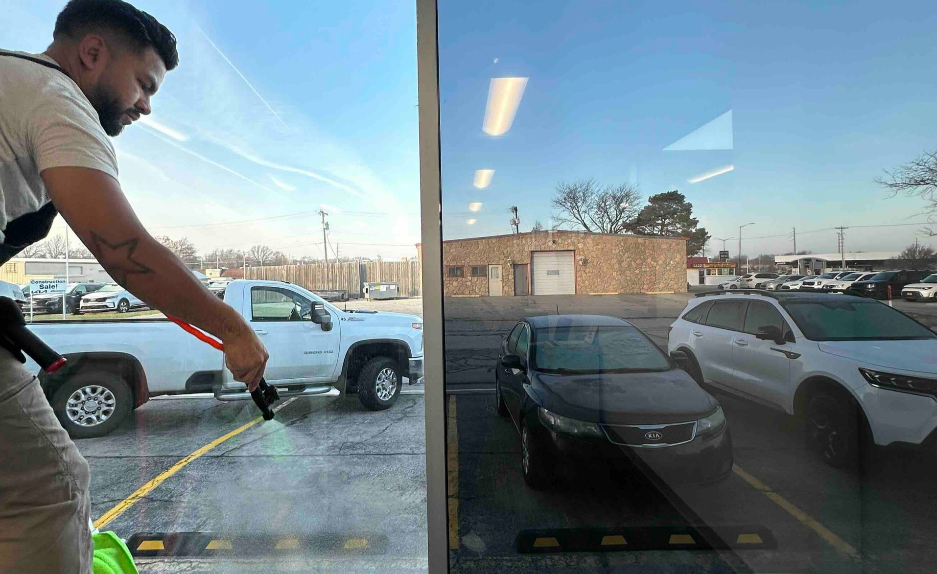 A person sprays a cleaner towards a truck parked in a parking lot, with cars visible in the reflection.