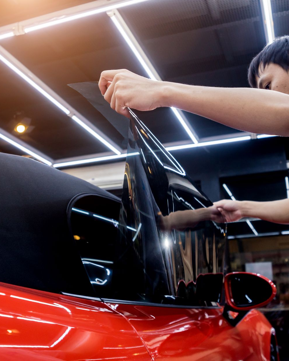 Man applying window tint to a red car window in a garage.
