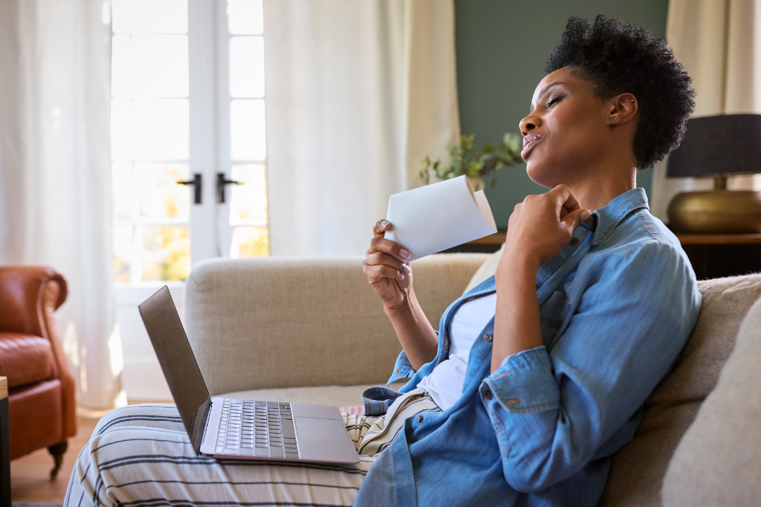 A woman is sitting on a couch with a laptop and fanning herself with an envelope.