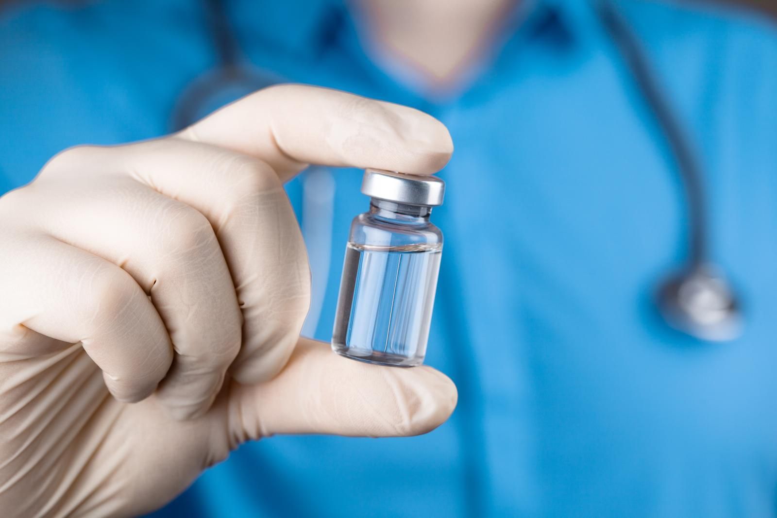Gloved hand holding a small peptide vial in front of a blue medical uniform and stethoscope
