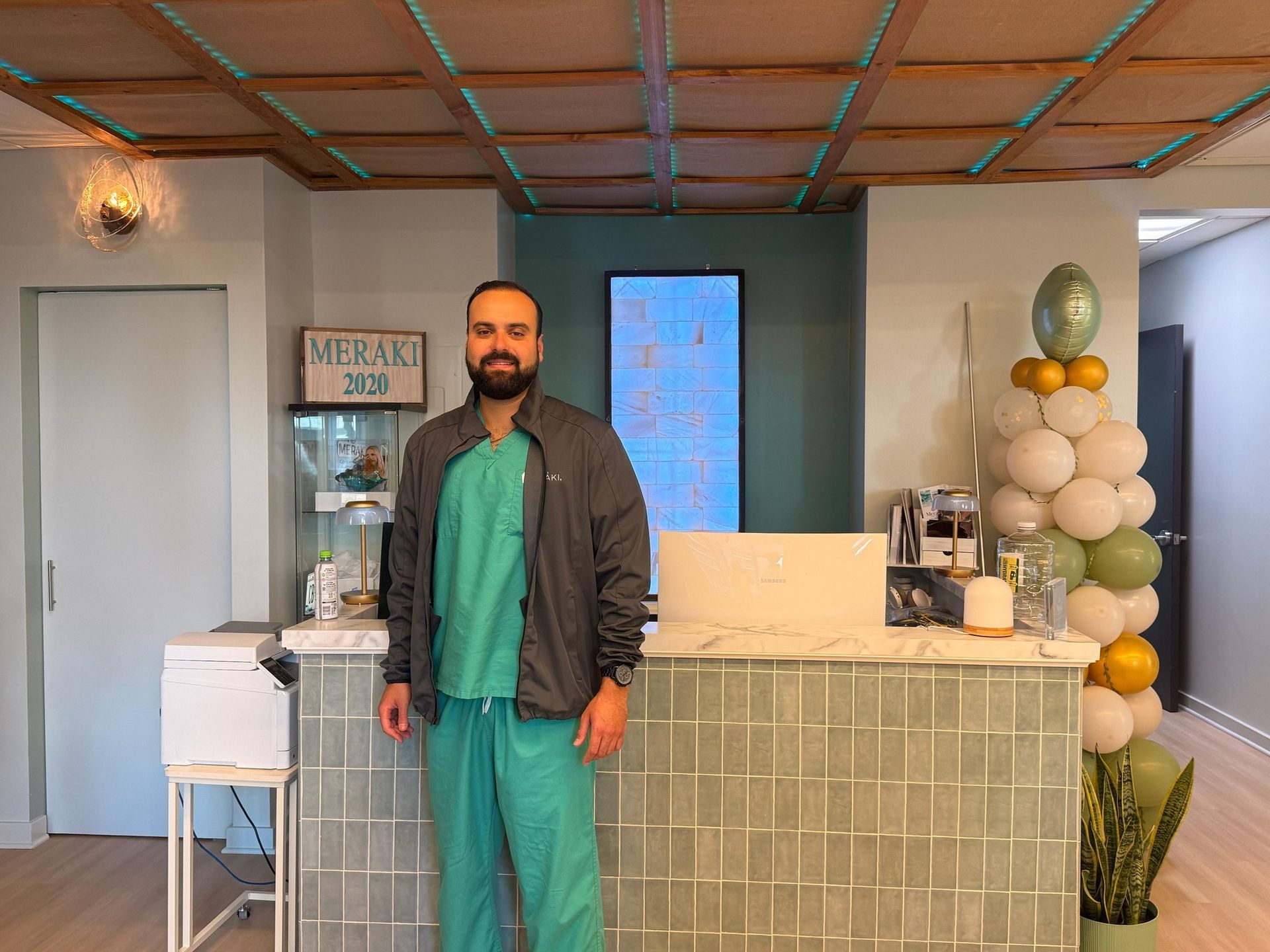 Man in green scrubs and a jacket stands at a reception desk with a balloon arch.