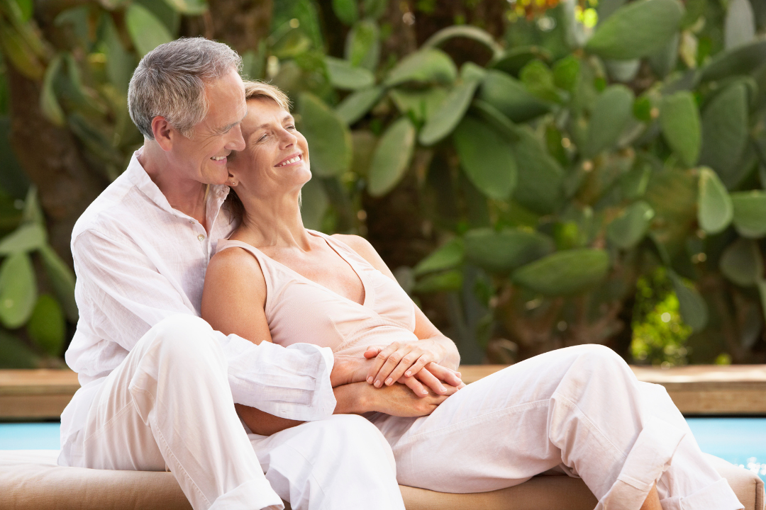 Couple in white clothing relaxing by a pool, smiling with greenery behind them