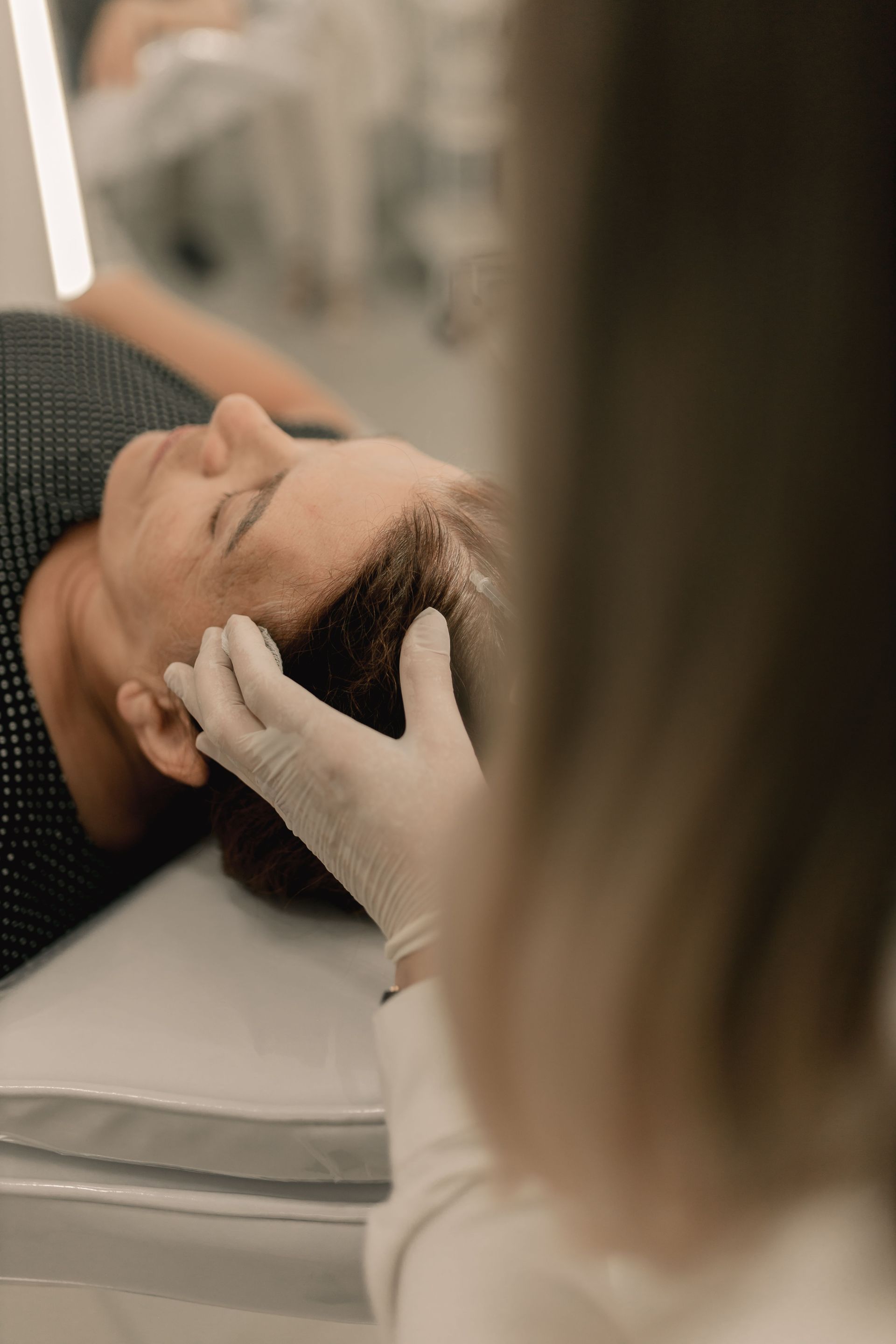 Gloved hands gently apply a hair restoration treatment to a person lying on a spa bed