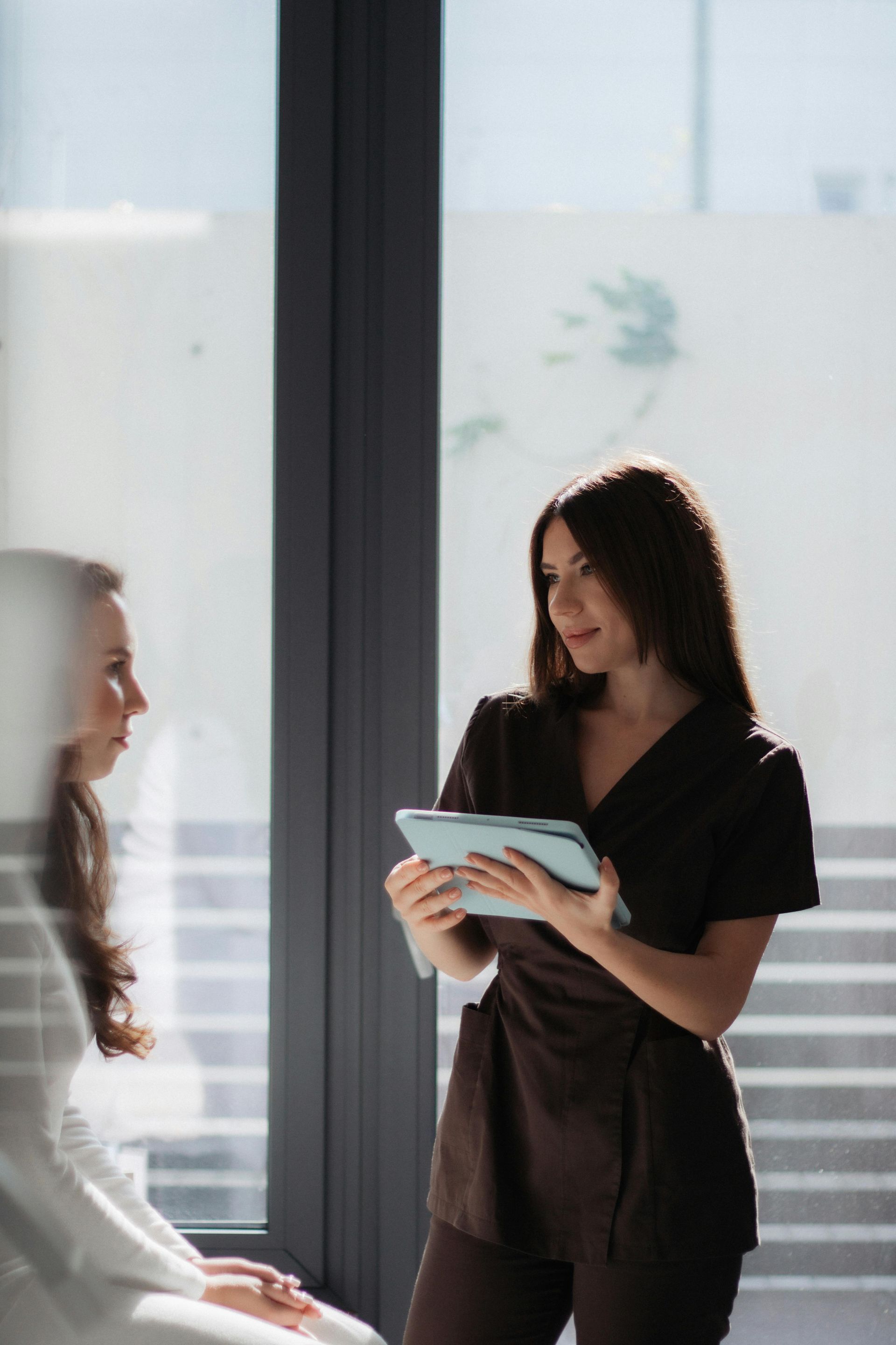 Two women in black business attire talking by a sunlit office window, one holding a notebook