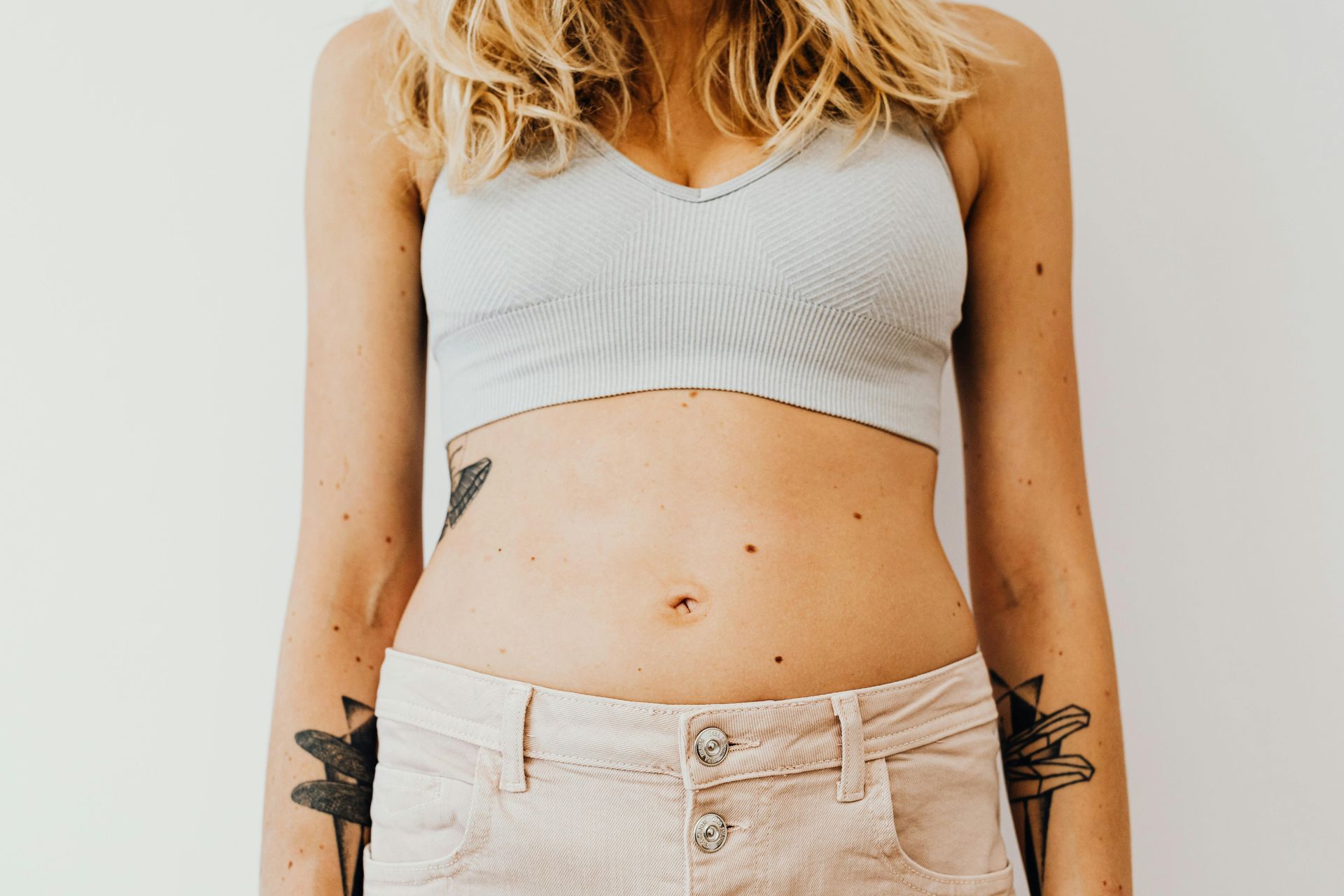 Woman’s torso in a gray crop top and beige pants with a tattoo visible on her side