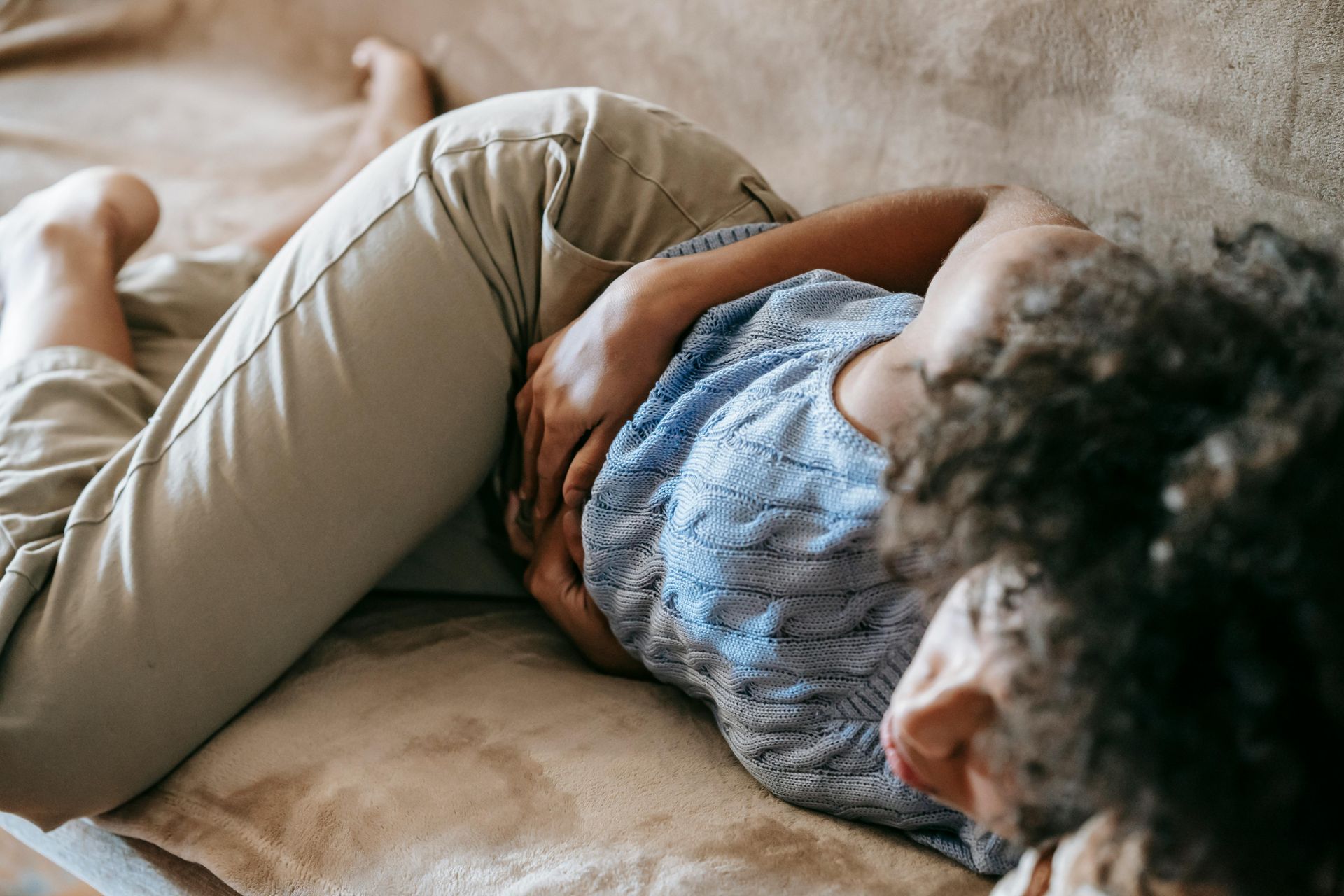 A person with curly hair lies on their side on a couch, holding their abdomen as if in pain.