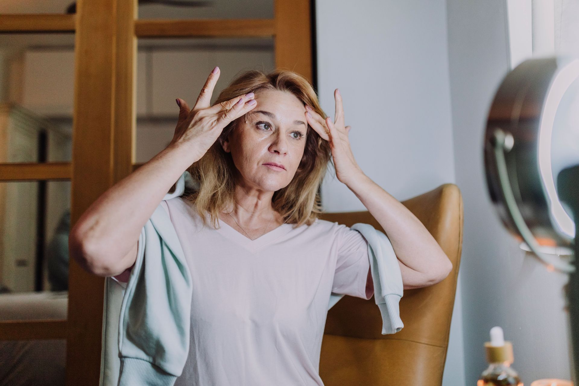 Woman in a white shirt touching her temples in a bedroom mirror, looking thoughtful or stressed