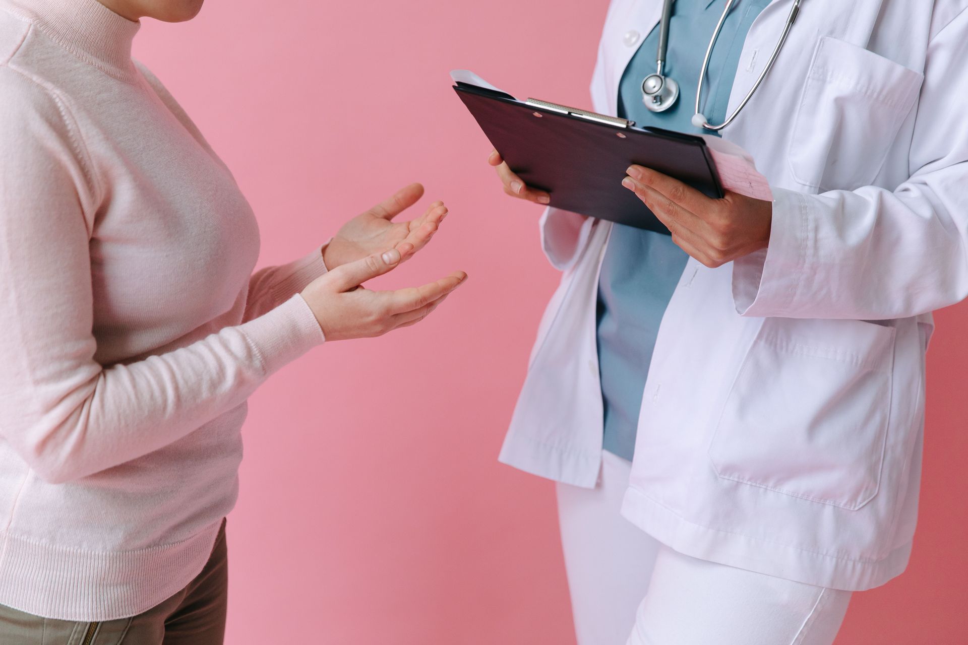 Doctor in white coat speaking with patient against pink background, holding a clipboard.