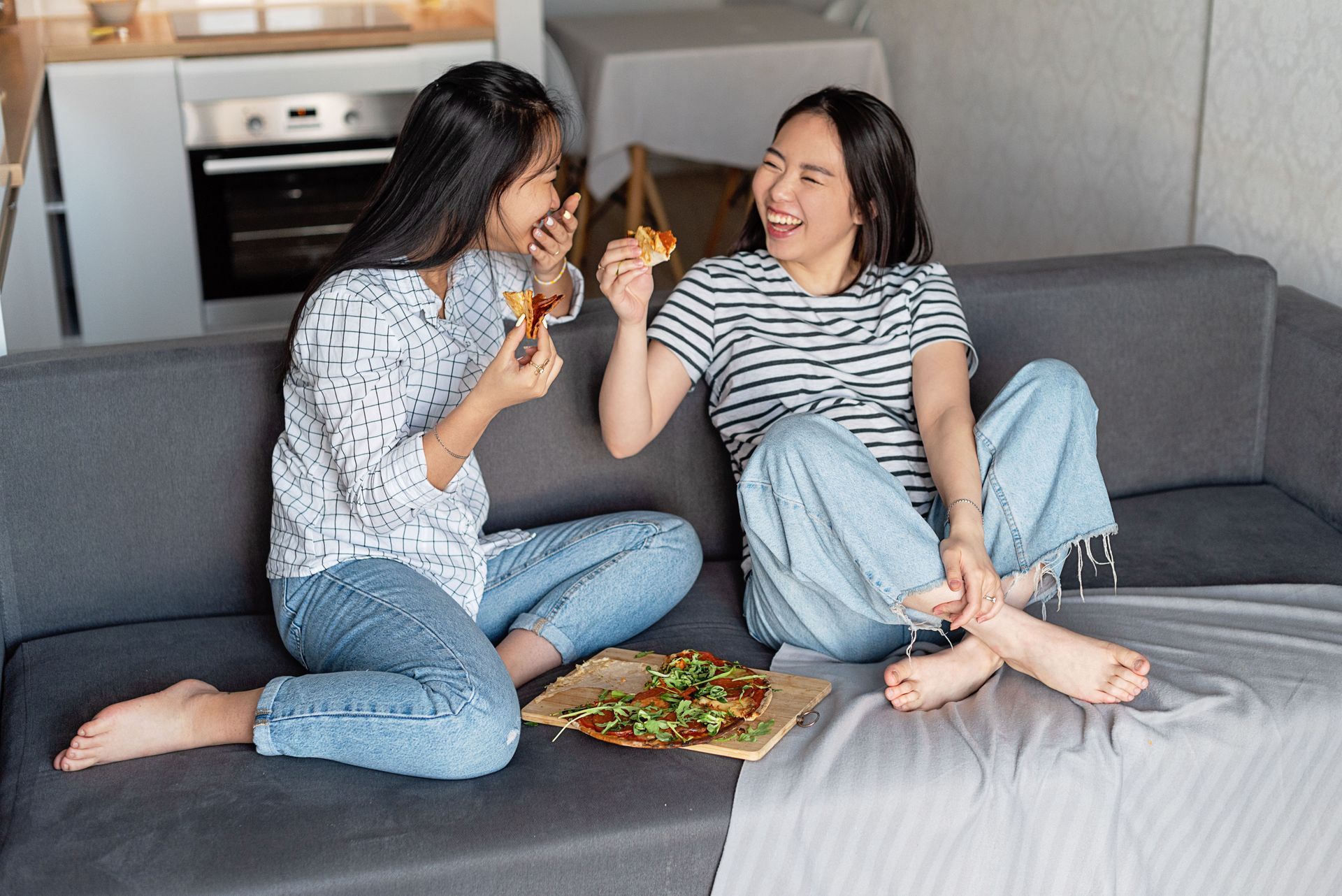 Two people sharing pizza on a gray couch, smiling in a living room.